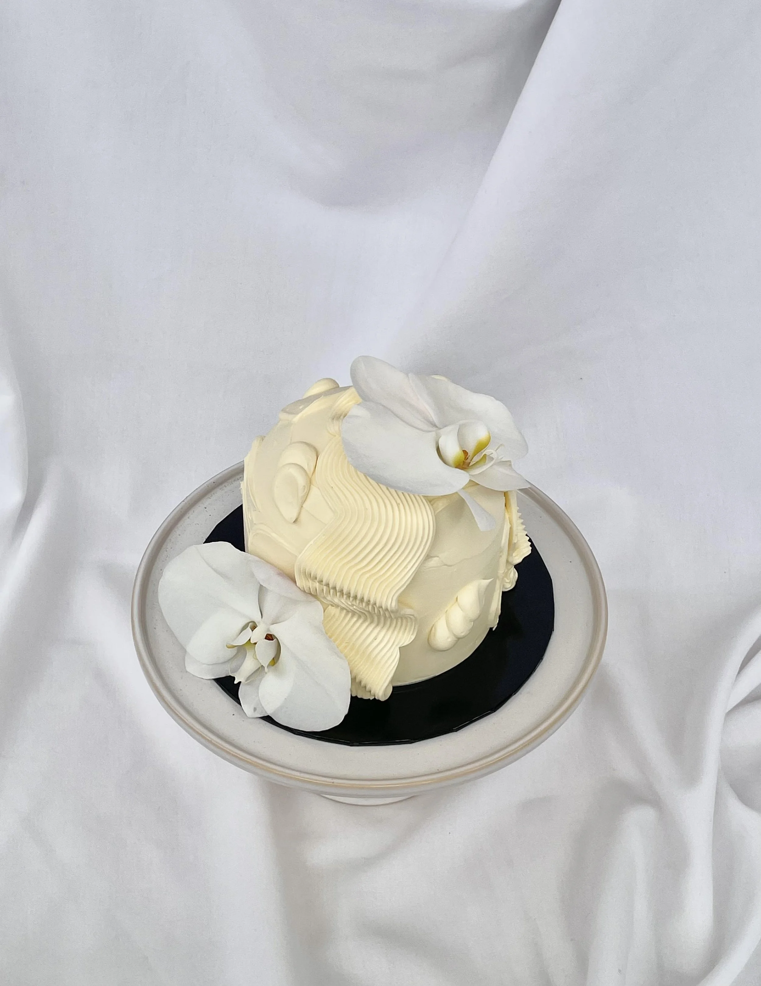 A small dome shaped cake topped with white orchid flowers, with a decorative cream swirl on top, served on a black and white plate.