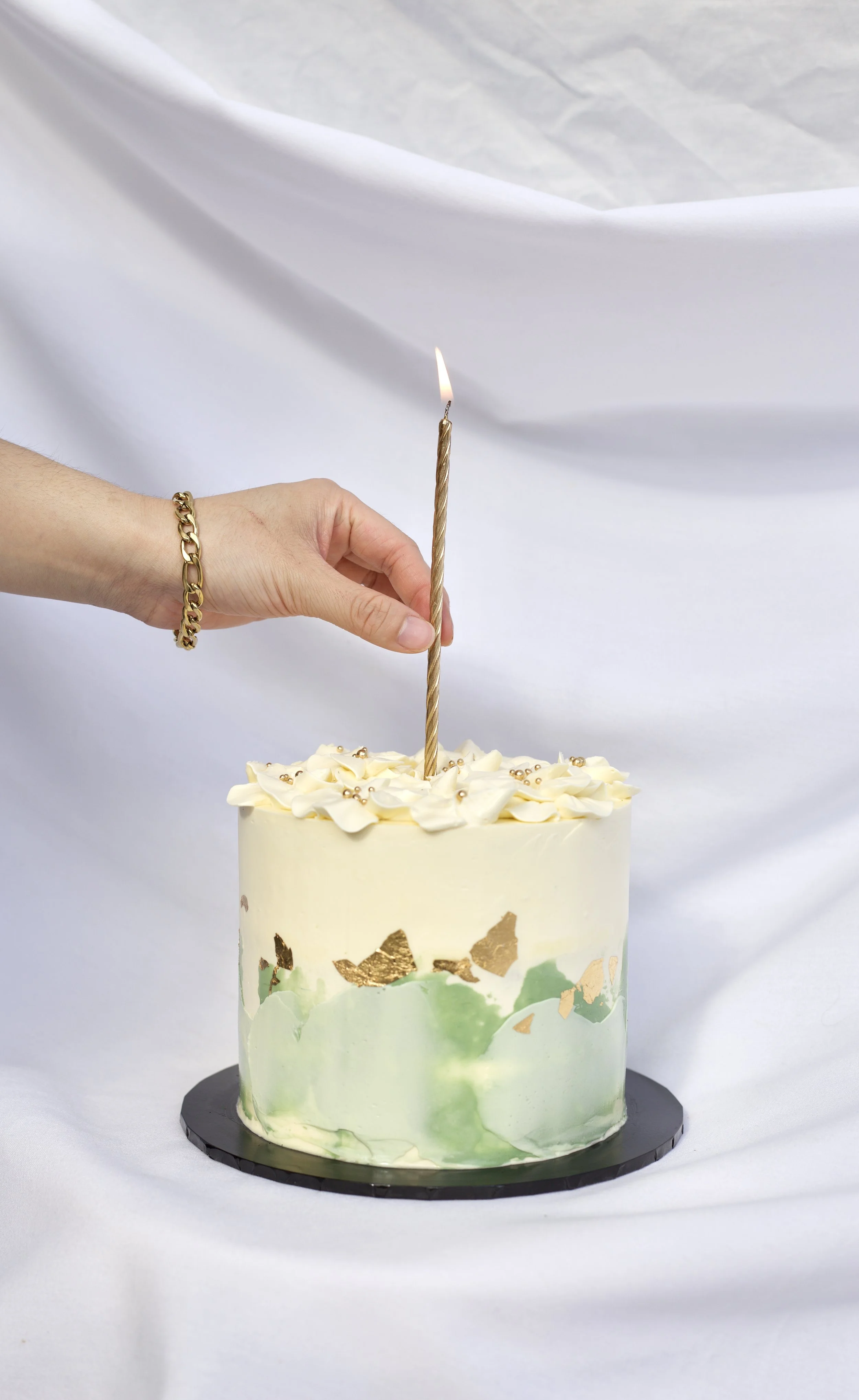 A person with a gold bracelet is lighting a tall, twisted gold birthday candle on a white frosted cake with decorative white frosting flowers and gold leaf accents, placed on a black cake board against a white fabric backdrop.