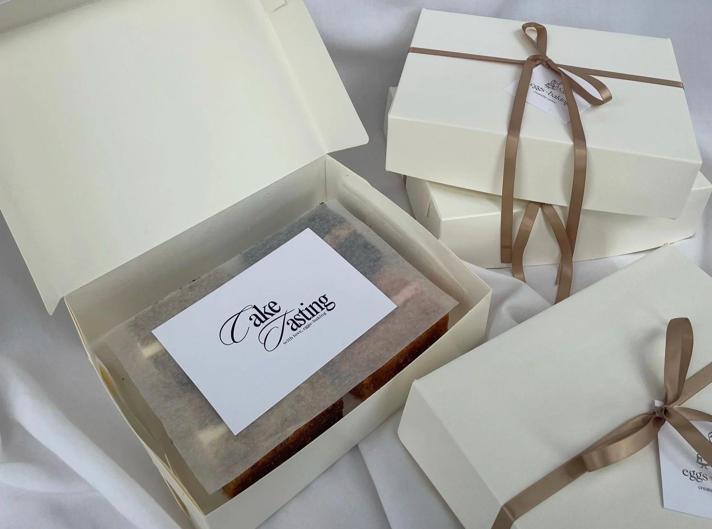 Cream-colored gift boxes with brown ribbons and tags, and a cake inside a cream-colored box labeled "Cake Tasting" with a website www.eggs-baking.com.
