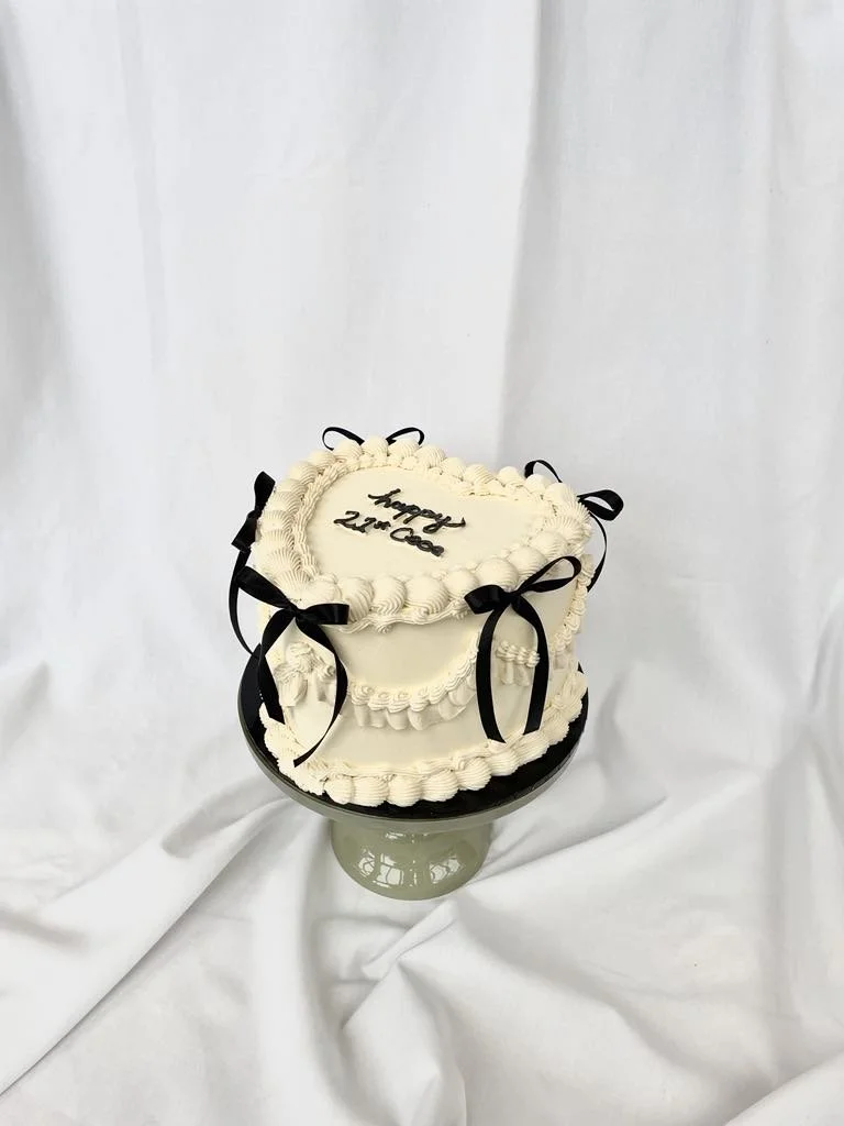 Heart-shaped birthday cake with white frosting, black ribbons, and black writing that says 'happy 22nd Coke' on top, on a cake stand with a white fabric backdrop.