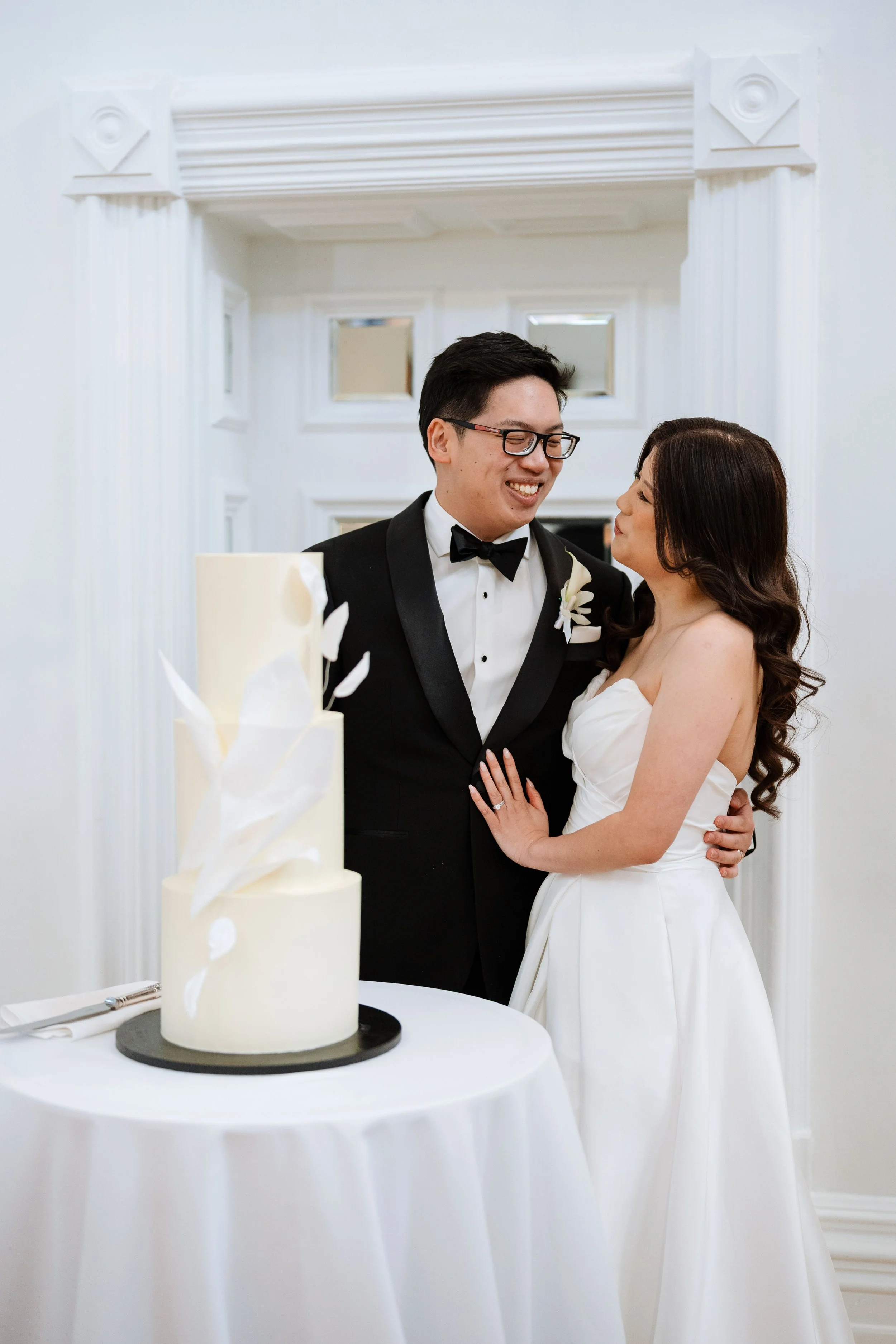 A newlywed couple dressed in wedding attire smiling and standing close together beside a wedding cake in a white decorated room.