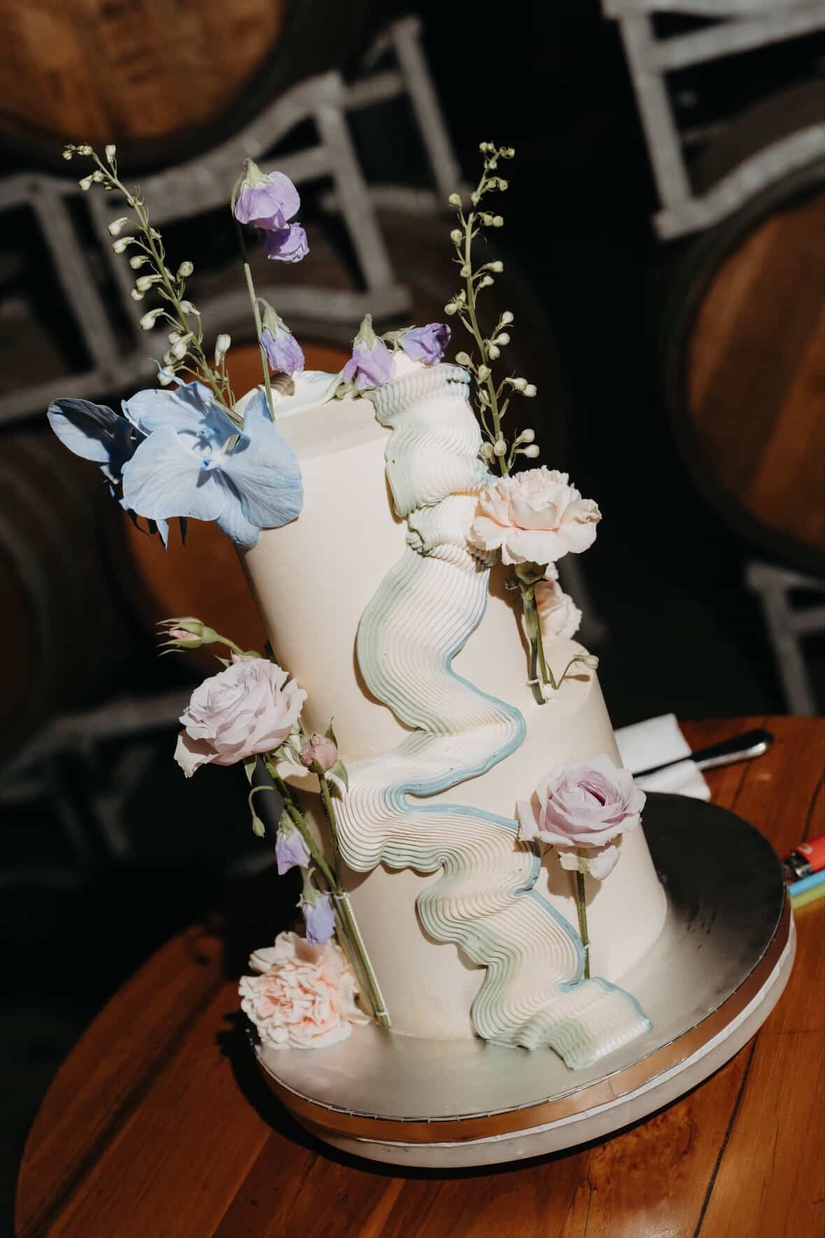 A tall, cylindrical white cake decorated with pastel-colored flowers and wavy designs, placed on a round metallic cake board on a wooden table.