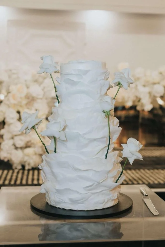 White crumpled paper-like sculpture with white paper flowers on green stems, displayed on a circular black base.