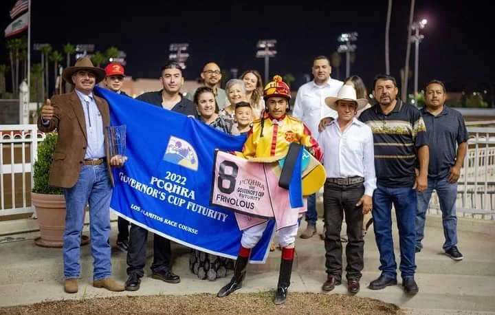 Group of people at a racetrack holding a blue flag and pink racing jersey, with a jockey in colorful racing attire in front.