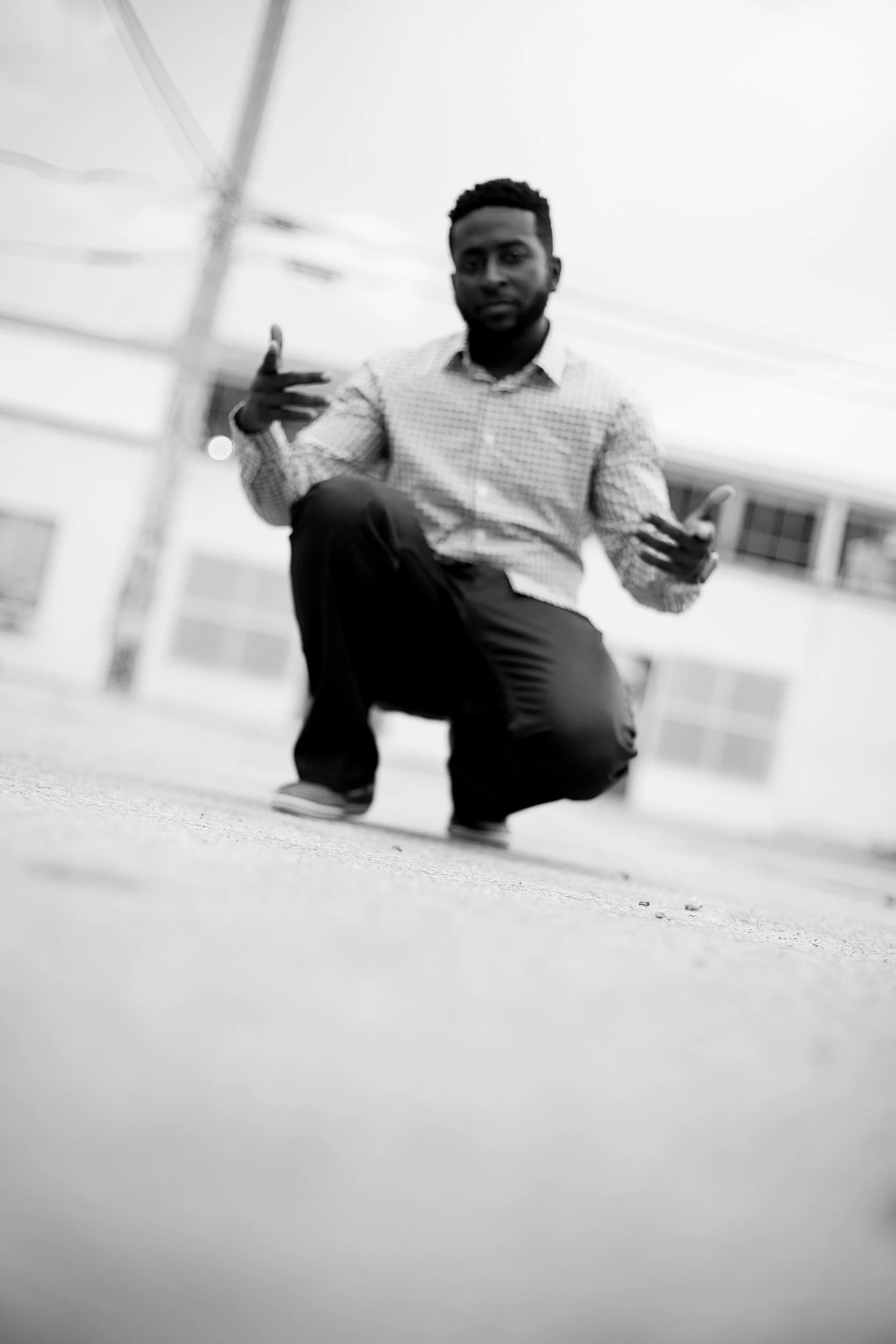 Herve Alce crouching on a street, making hand gestures, with power lines and buildings in the background, in black and white.