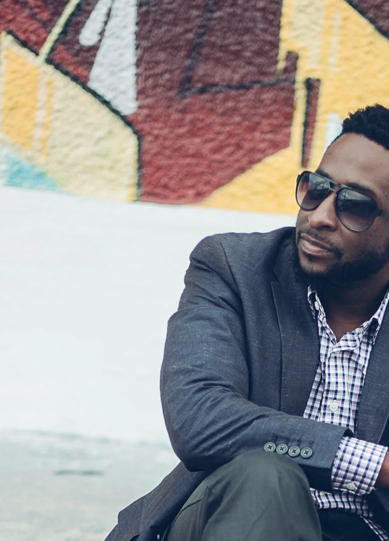 Herve Alce wearing sunglasses and a blazer sitting outdoors with a colorful mural in the background.