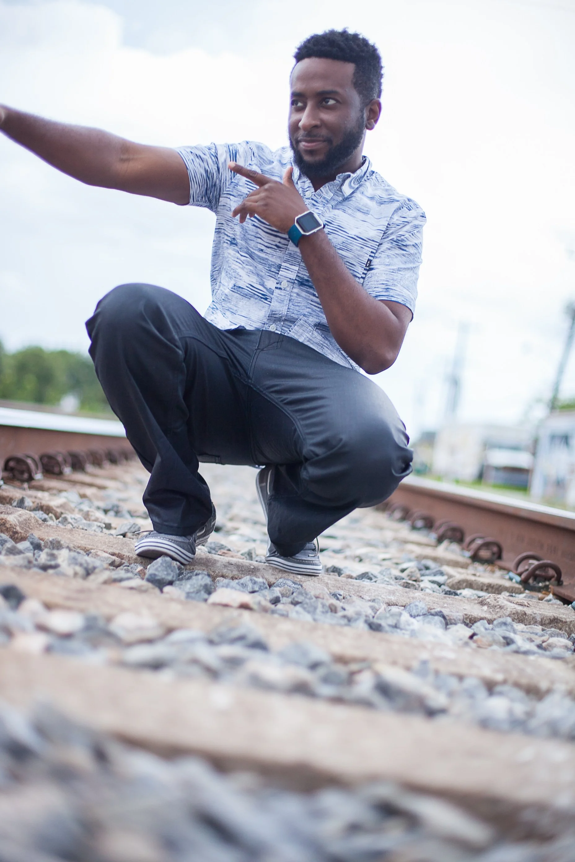 Here Alce squatting on a railroad track, pointing to the side, with a thoughtful expression, wearing a patterned short-sleeve shirt, black pants, and a smartwatch.