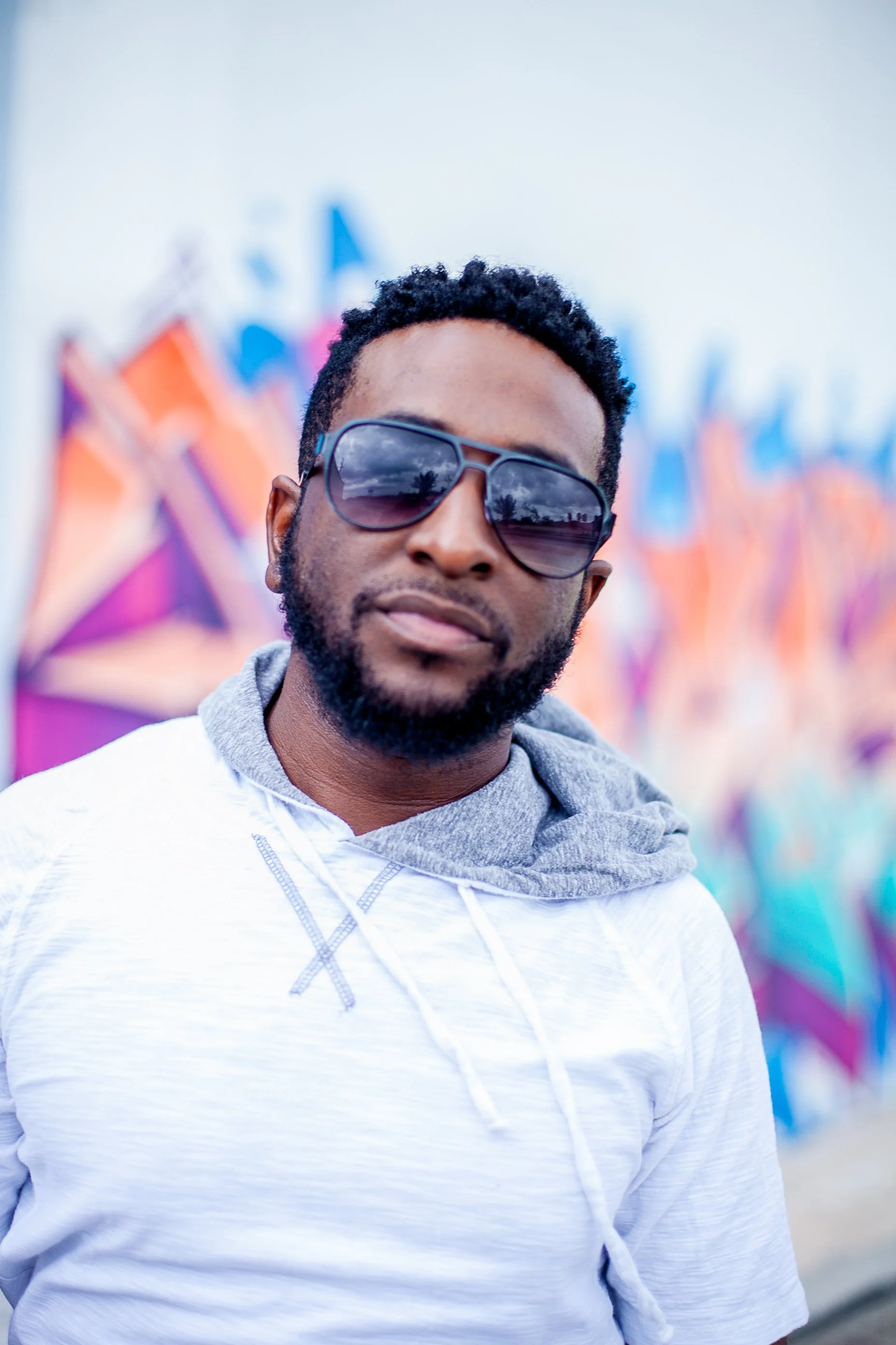 A man with a beard wearing sunglasses, a white hoodie, and a gray sweatshirt, standing in front of a colorful graffiti wall.
