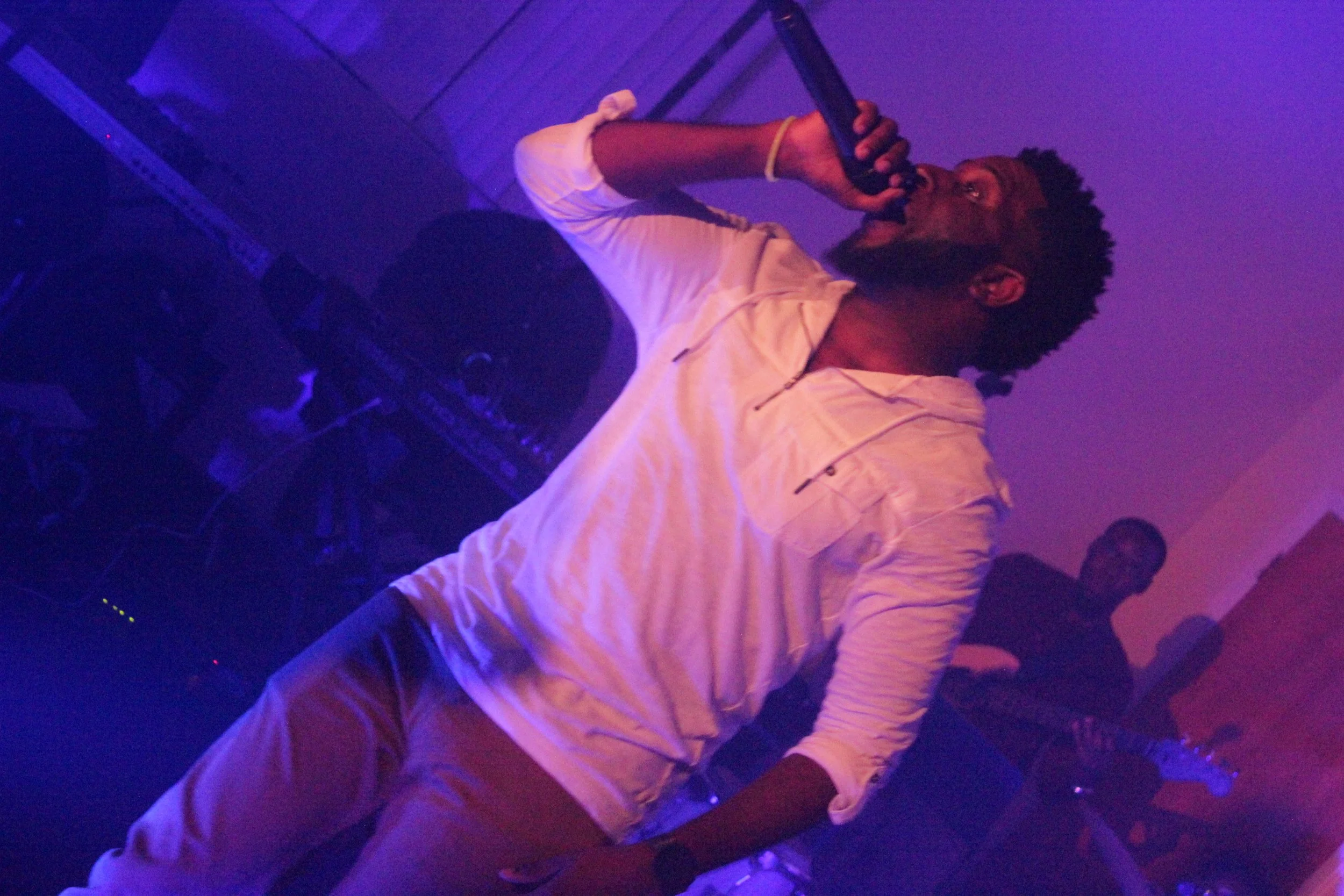 Herve Alce singing into a microphone on stage with purple and blue lighting, with a musician playing a bass guitar in the background.