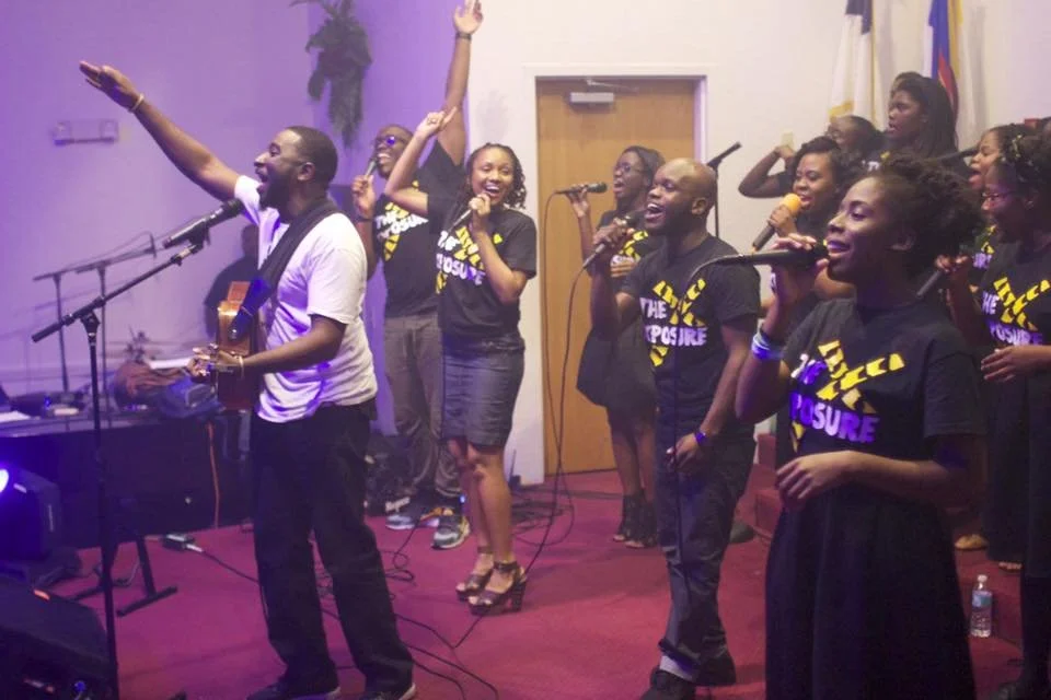 Group of people singing and Herve Alce playing guitar in a room with purple lighting, wearing matching black T-shirts.