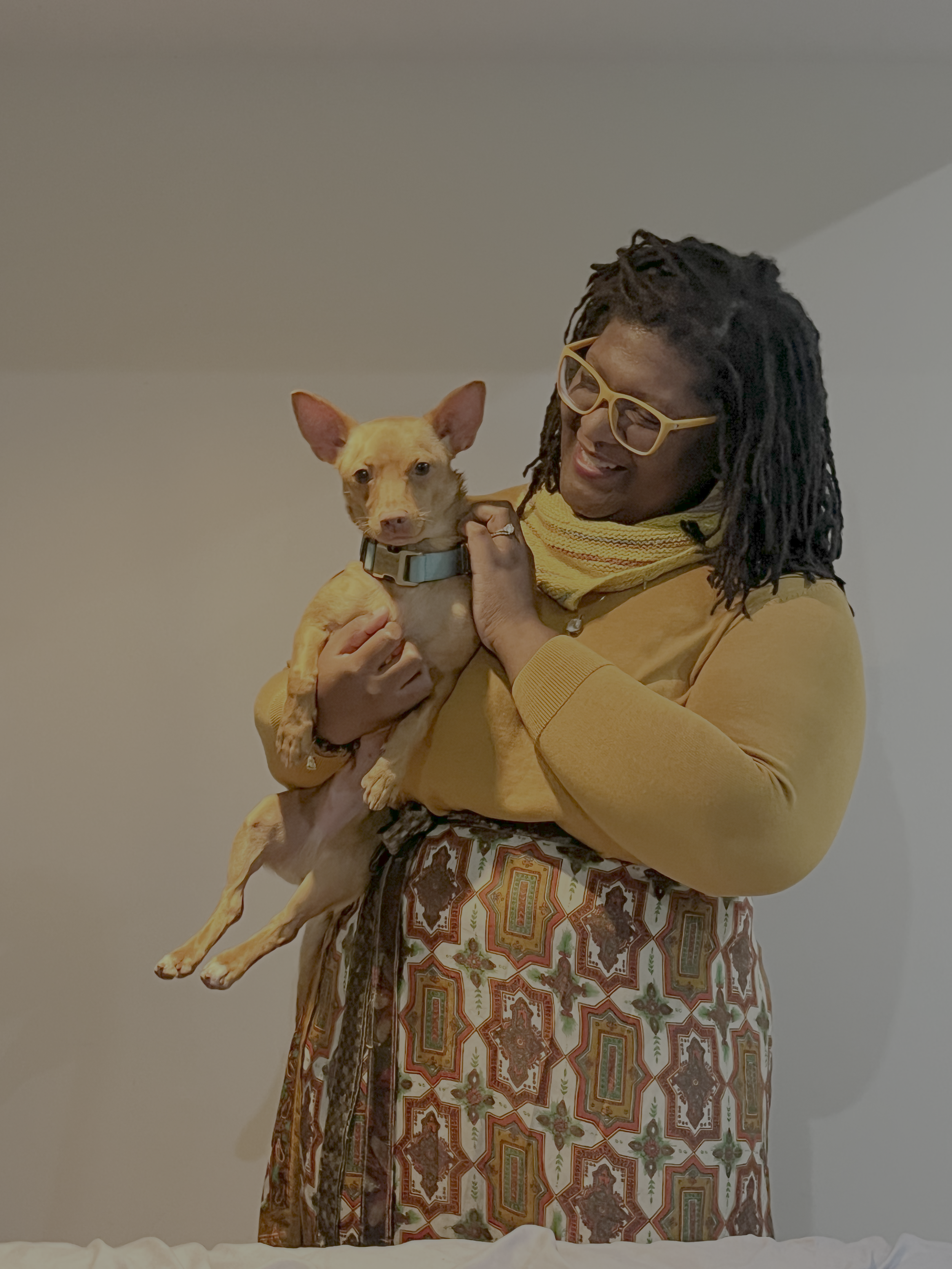 A woman with glasses and locks holding and smiling at a small tan dog with large ears, against a plain wall background.