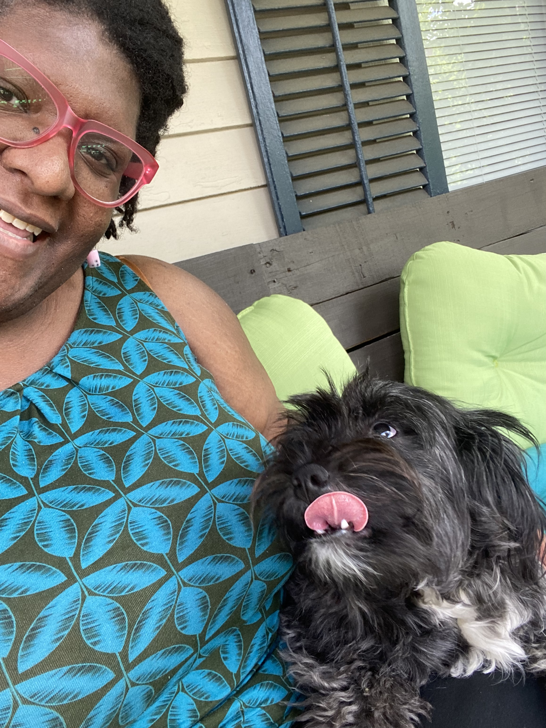 A woman with dark skin wearing pink glasses and a blue patterned sleeveless top, smiling while holding a black and white dog with its tongue out, sitting on a green cushion outside near a beige house with a window and shutter.