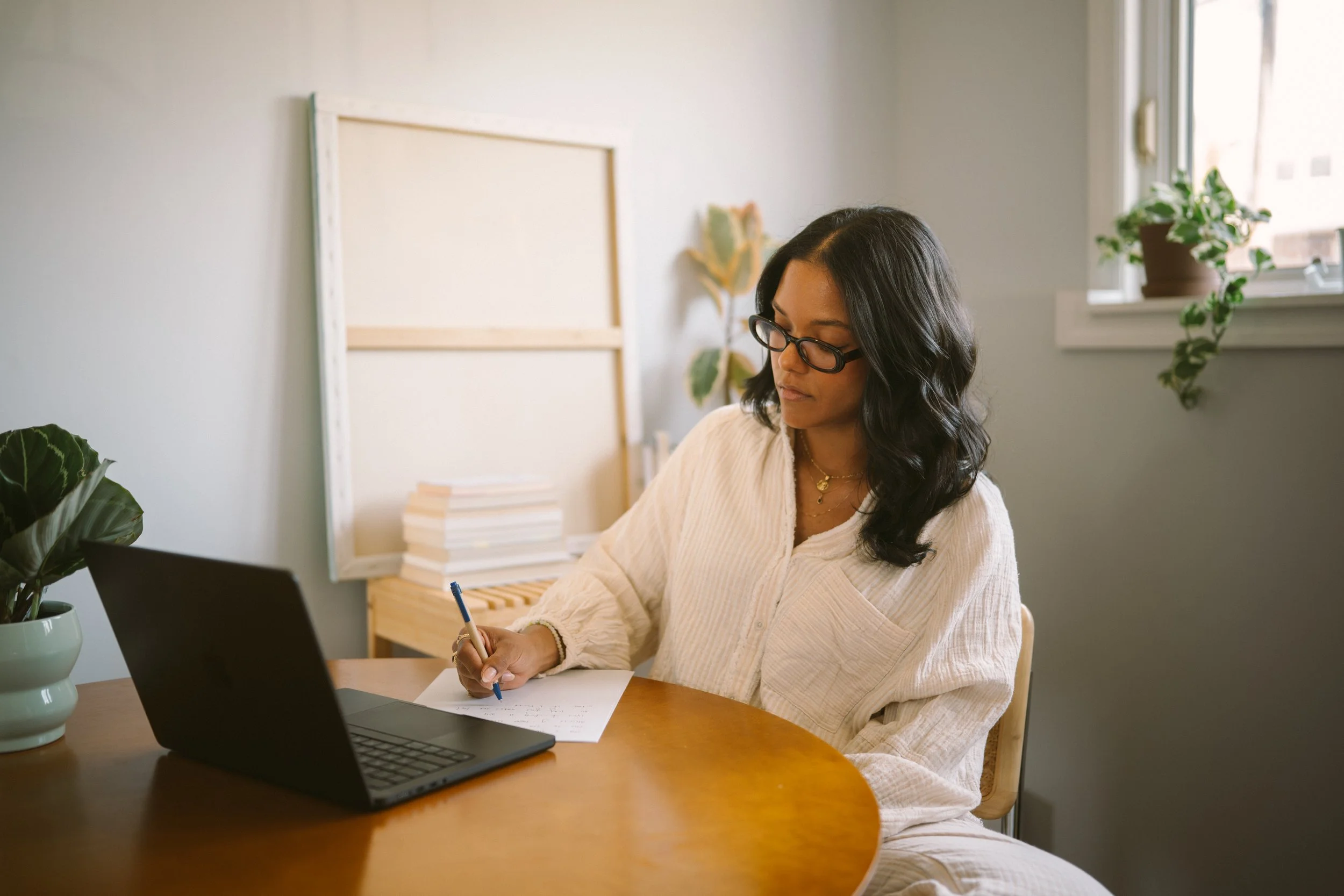 A woman with dark hair, glasses, and gold jewelry sitting at a wooden table, writing on paper with a blue pen, with a laptop, houseplants, and a blank corkboard in the background.