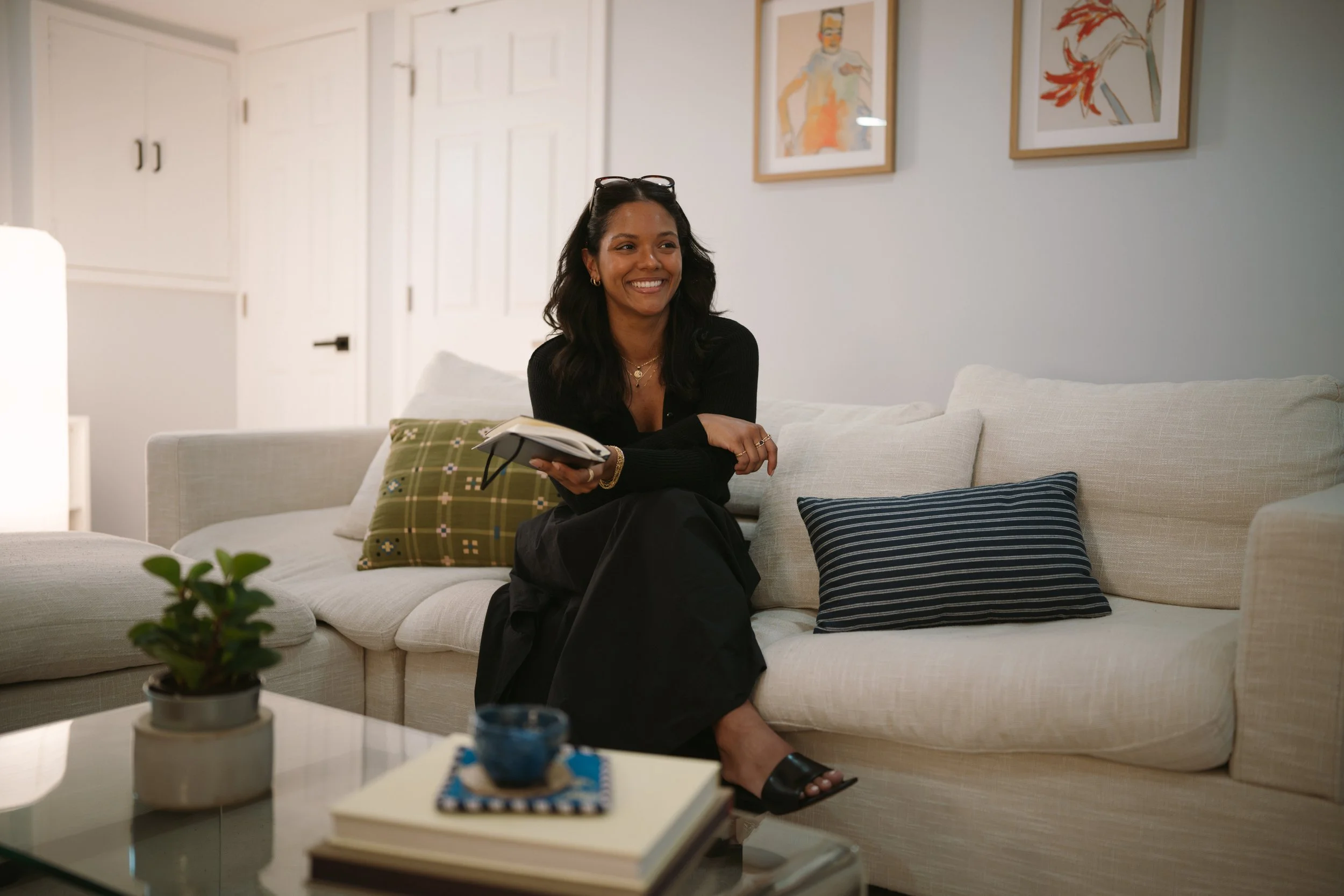 A woman with dark hair, gold jewelry, glasses on her head, wearing a black top and black pants, is smiling and sitting on a cream-colored sofa in a well-lit living room. She holds a notepad or book. Behind her, there are framed watercolor portraits on the wall. The sofa has striped and patterned pillows. A small plant is on the coffee table, and a blue decorative item is also visible.