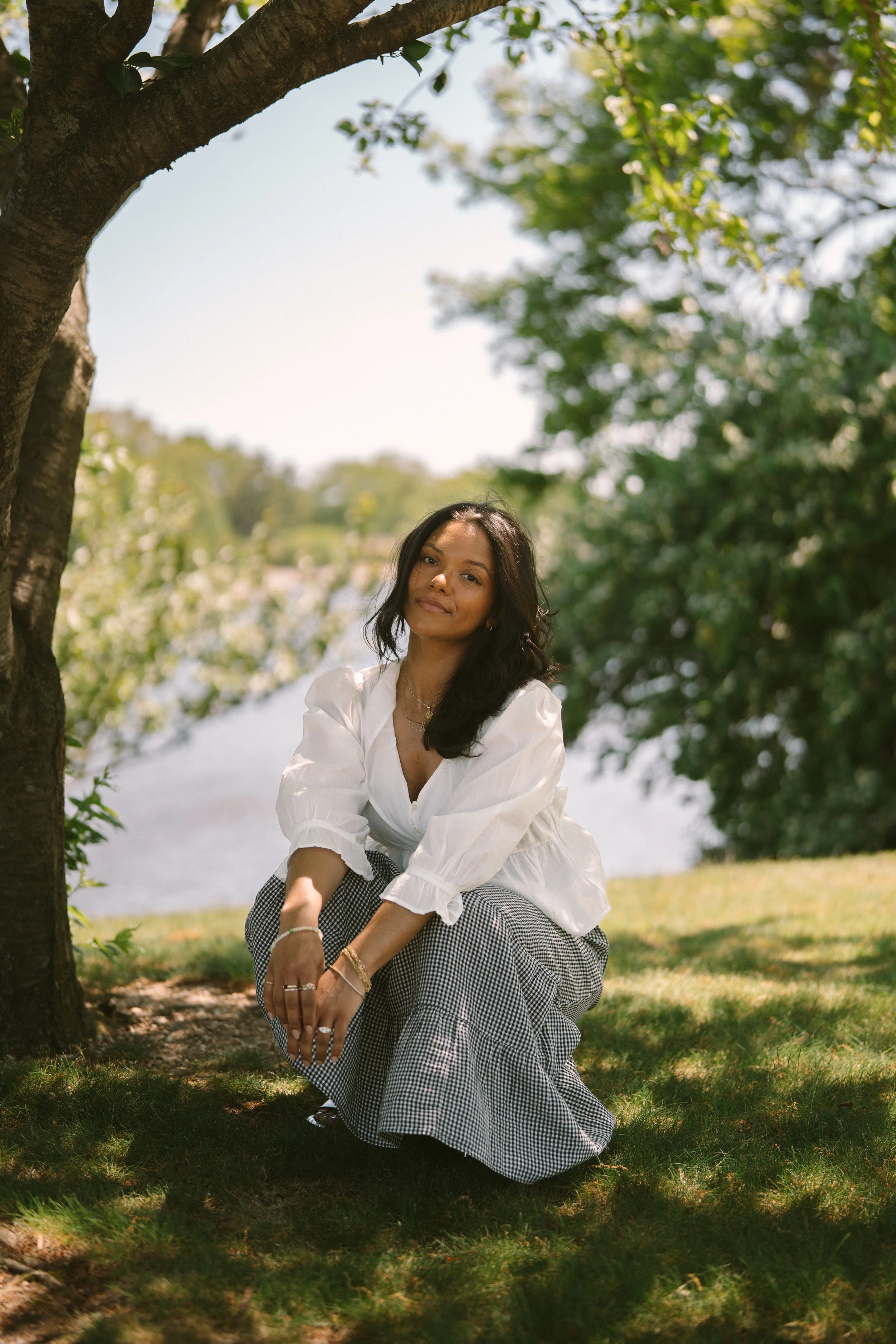 A woman crouching on the grass near a tree, smiling, with a lake and green trees in the background, on a sunny day.