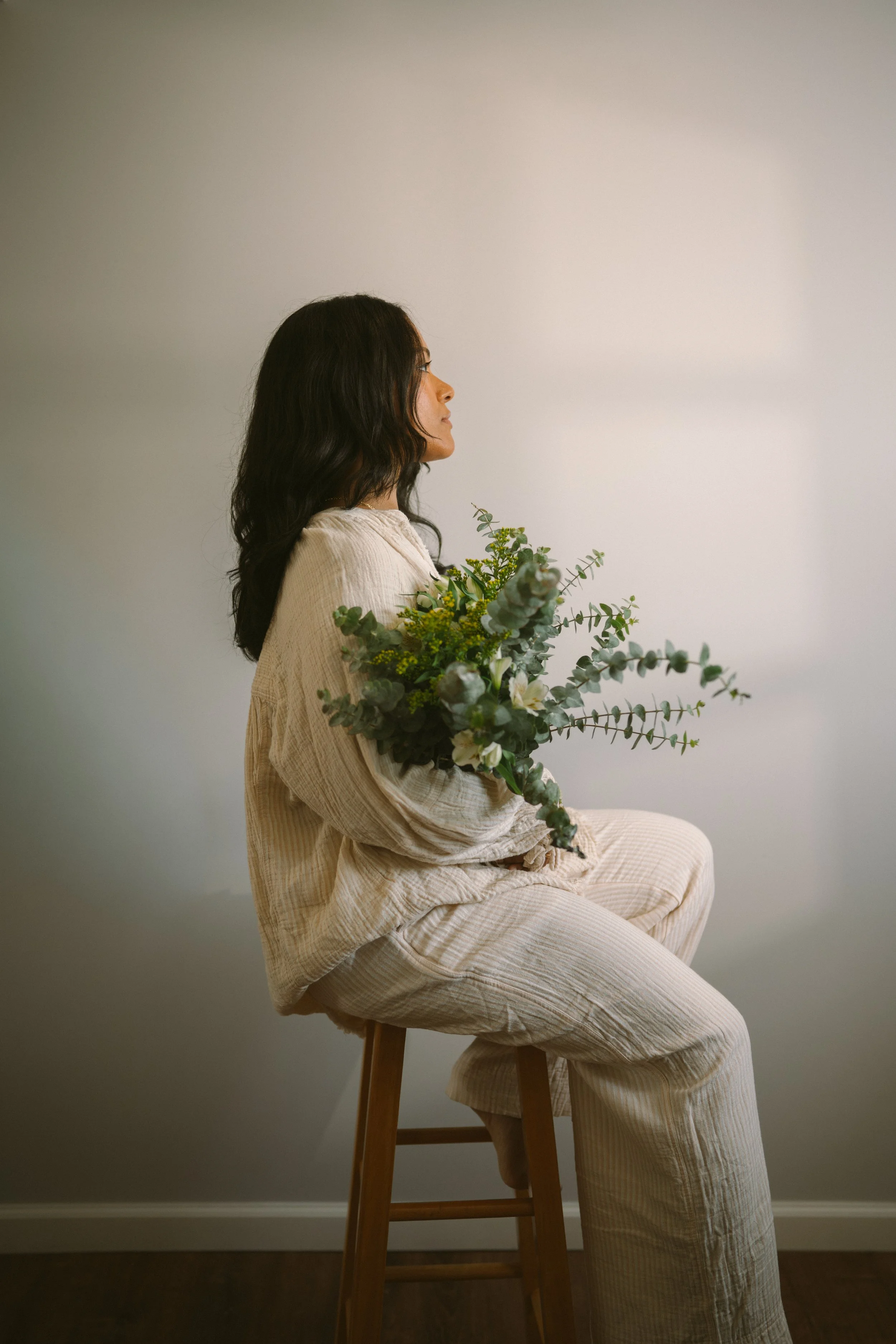 A woman with long dark wavy hair sitting on a wooden stool, holding a bouquet of eucalyptus and white flowers, wearing a beige loose-fitting outfit, against a plain white wall.