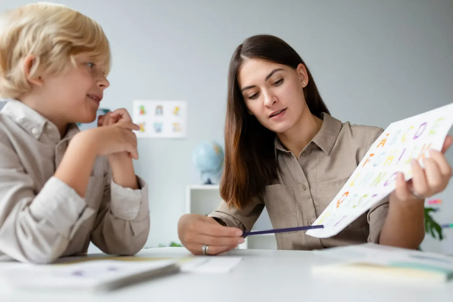 A woman and a young boy sitting at a table looking at a colorful sheet of paper, in a classroom or office setting.