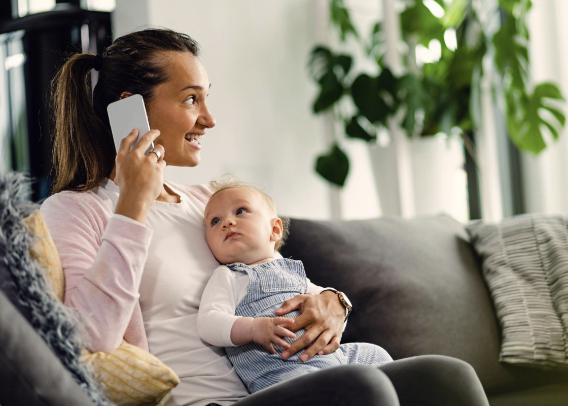 A woman sitting on a gray couch, talking on a cell phone, while holding a young child on her lap who is looking up at her. There are houseplants and pillows in the background.