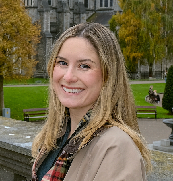 A woman with blonde hair smiling outdoors in a park with trees, benches, and an old building in the background.