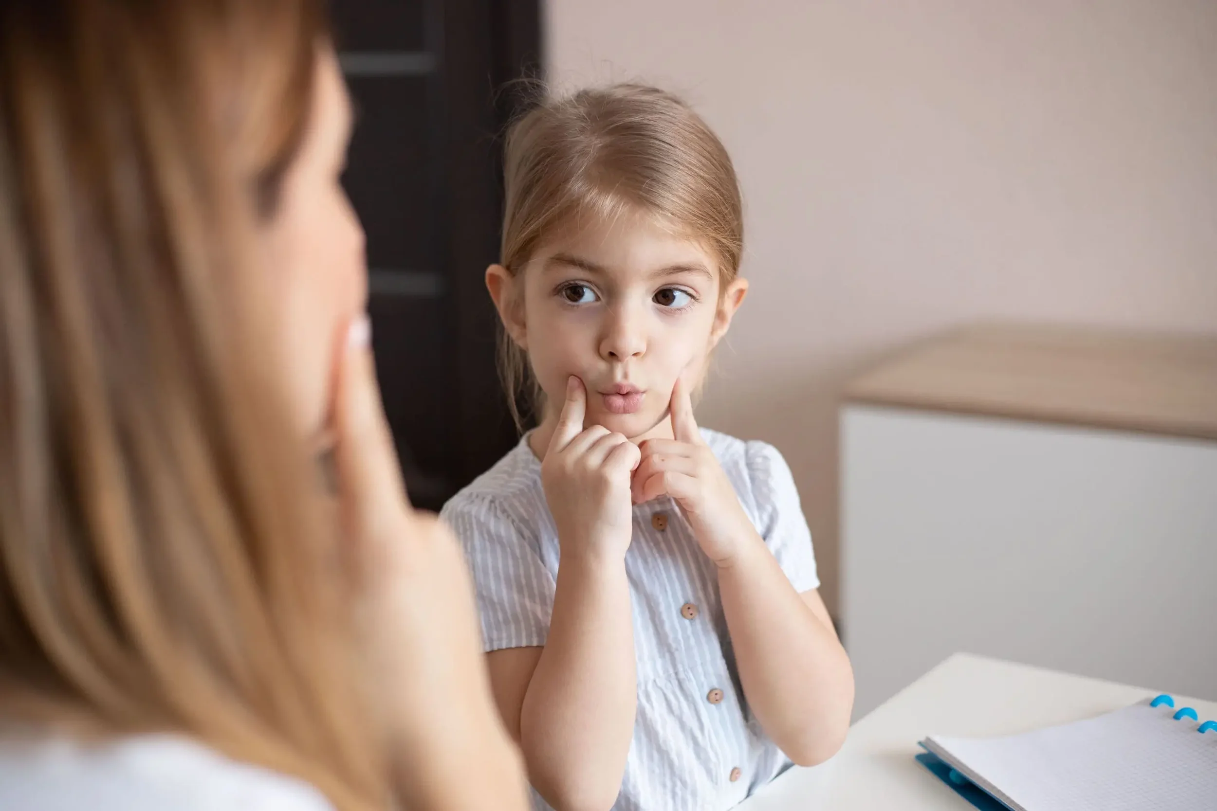 A young girl sitting at a table, with a notebook in front of her, making a silly face by pressing her cheeks with her fingers while looking at an adult, who is slightly out of focus.