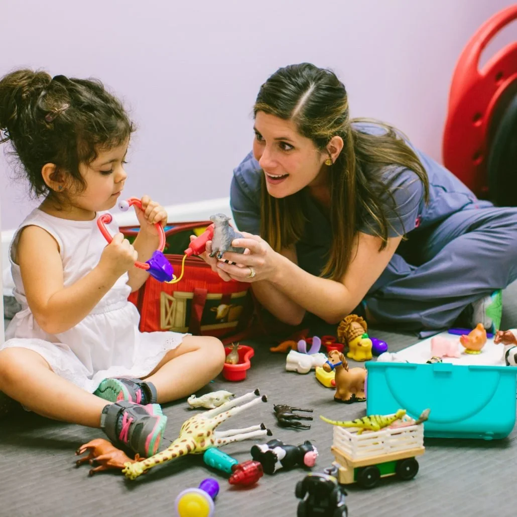 A woman and a young girl playing with toy animals and figurines during a pretend play session on the floor.
