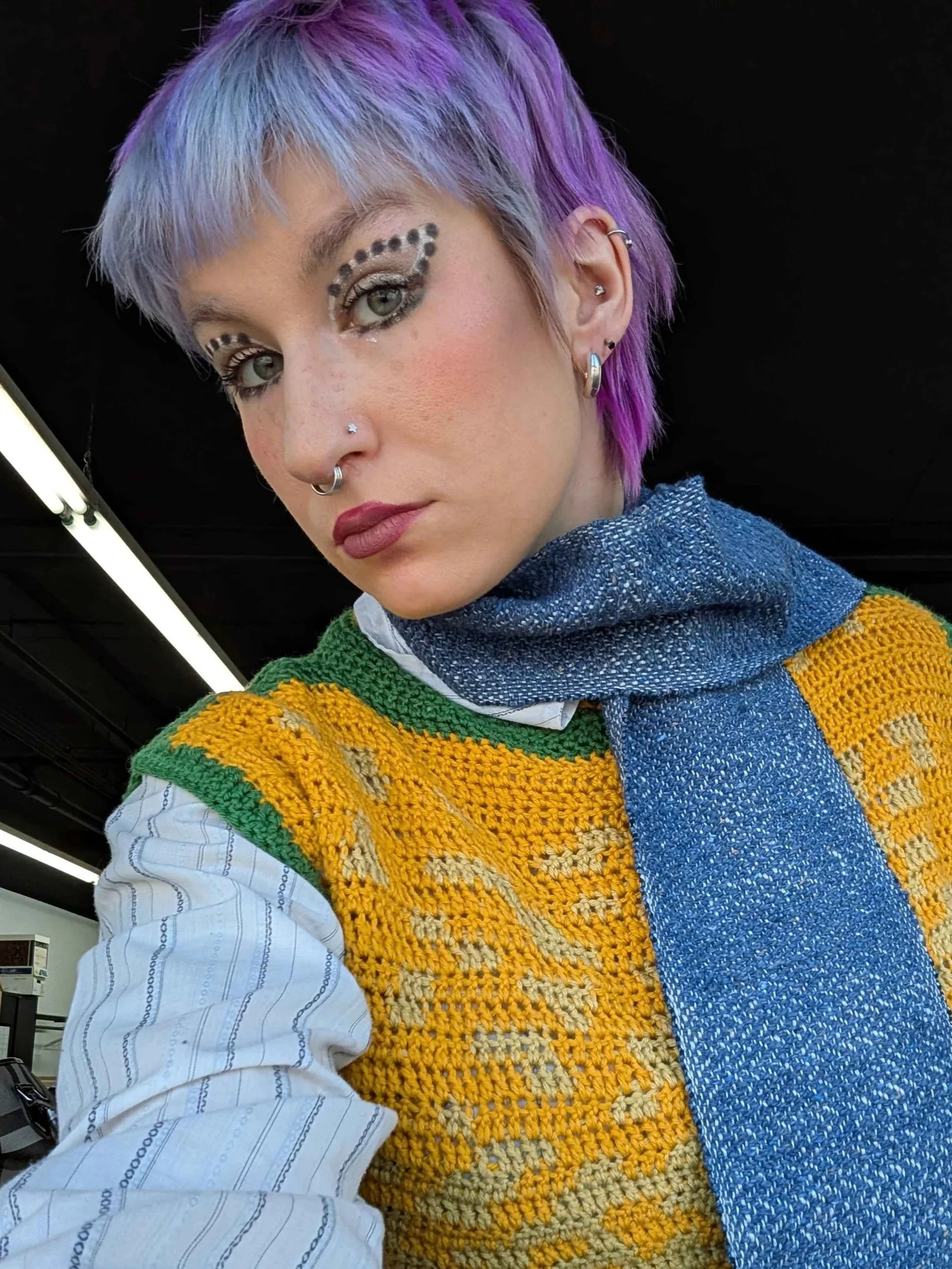 A young woman with light purple and pink hair, wearing a blue scarf, yellow and green knitted vest over a white shirt with thin blue stripes, and multiple earrings. She has makeup with eye designs and facial piercings.