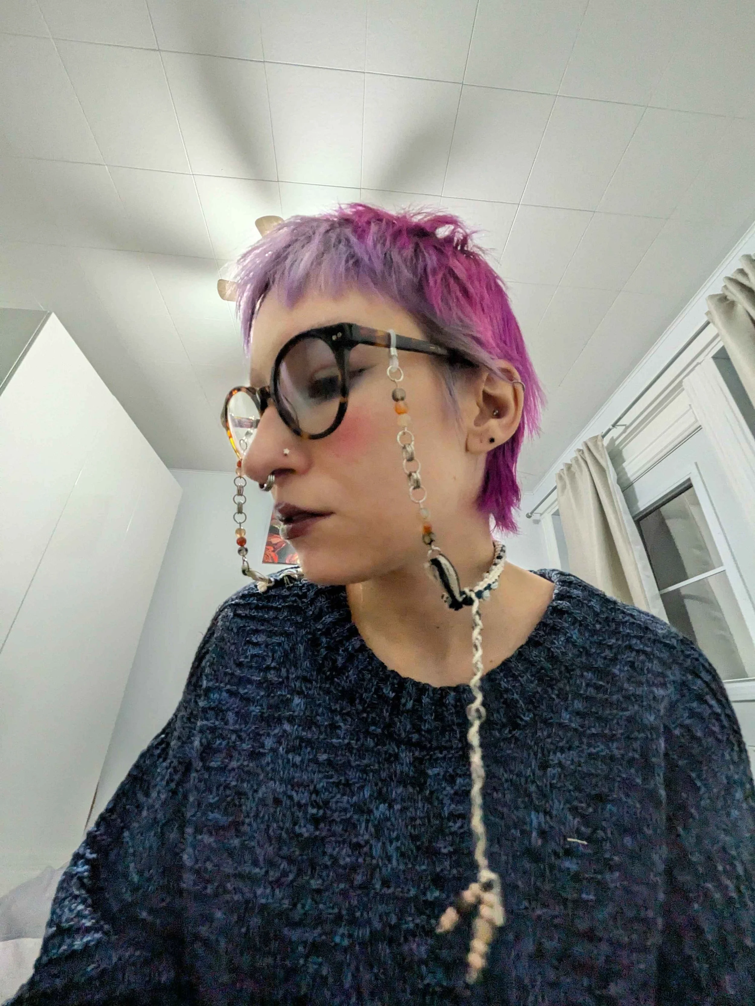 A person with pink and purple short hair, wearing black glasses with a chain, a septum piercing, and dark lipstick, sitting indoors with a white window with curtains in the background.