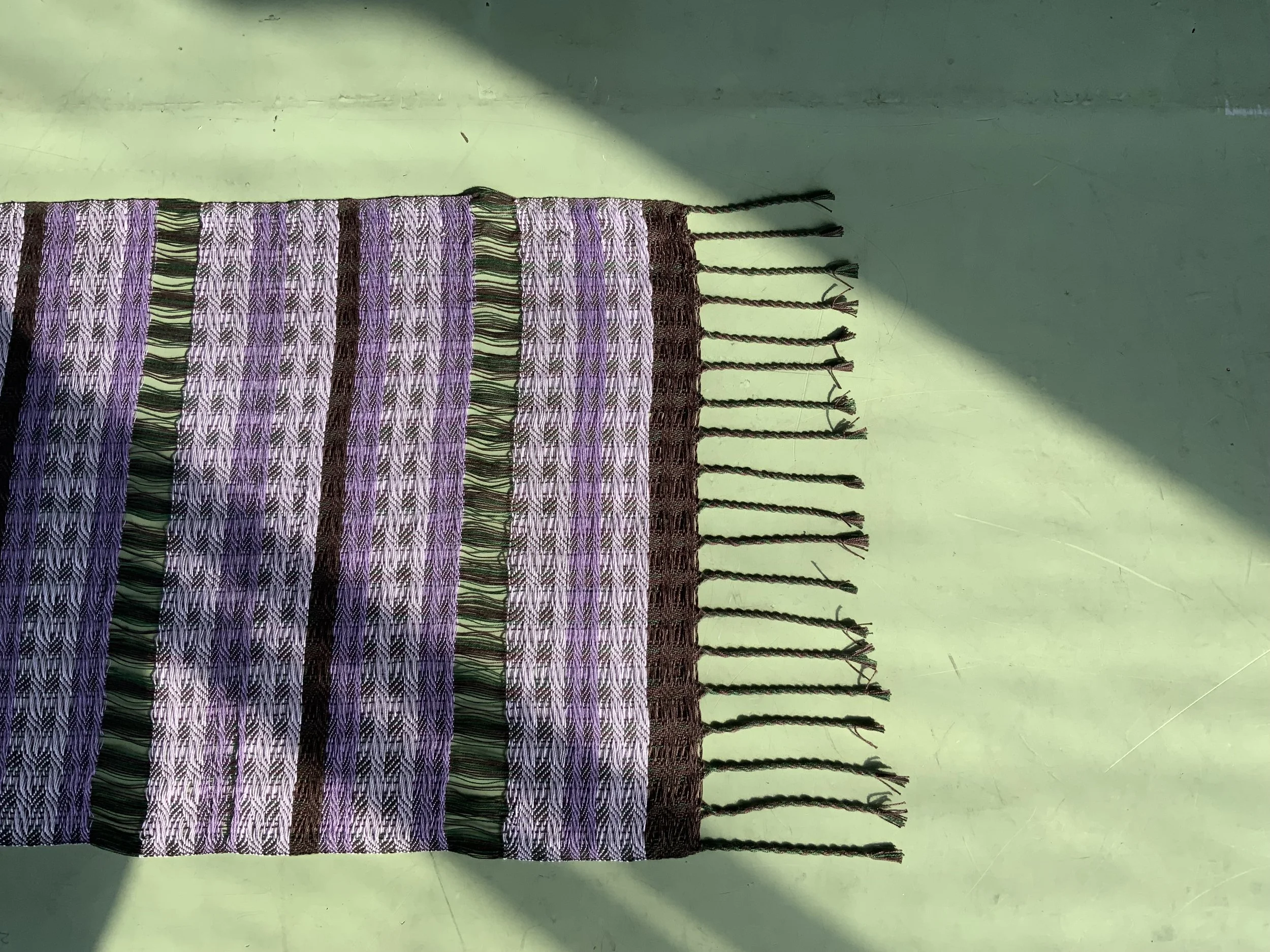 Close-up of a woven textile with purple, green, black, and white stripes, laid on a light green surface with a shadow cast over part of it.