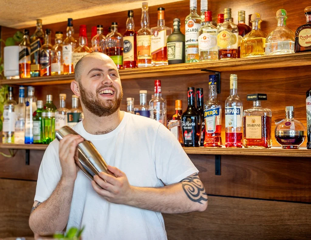 A bartender holding a cocktail shaker, smiling in front of a wooden shelf filled with various bottles of alcohol.