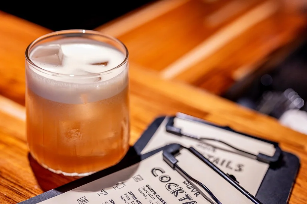 A glass of beer with a foamy head on a wooden table next to a clipboard with a music event flyer