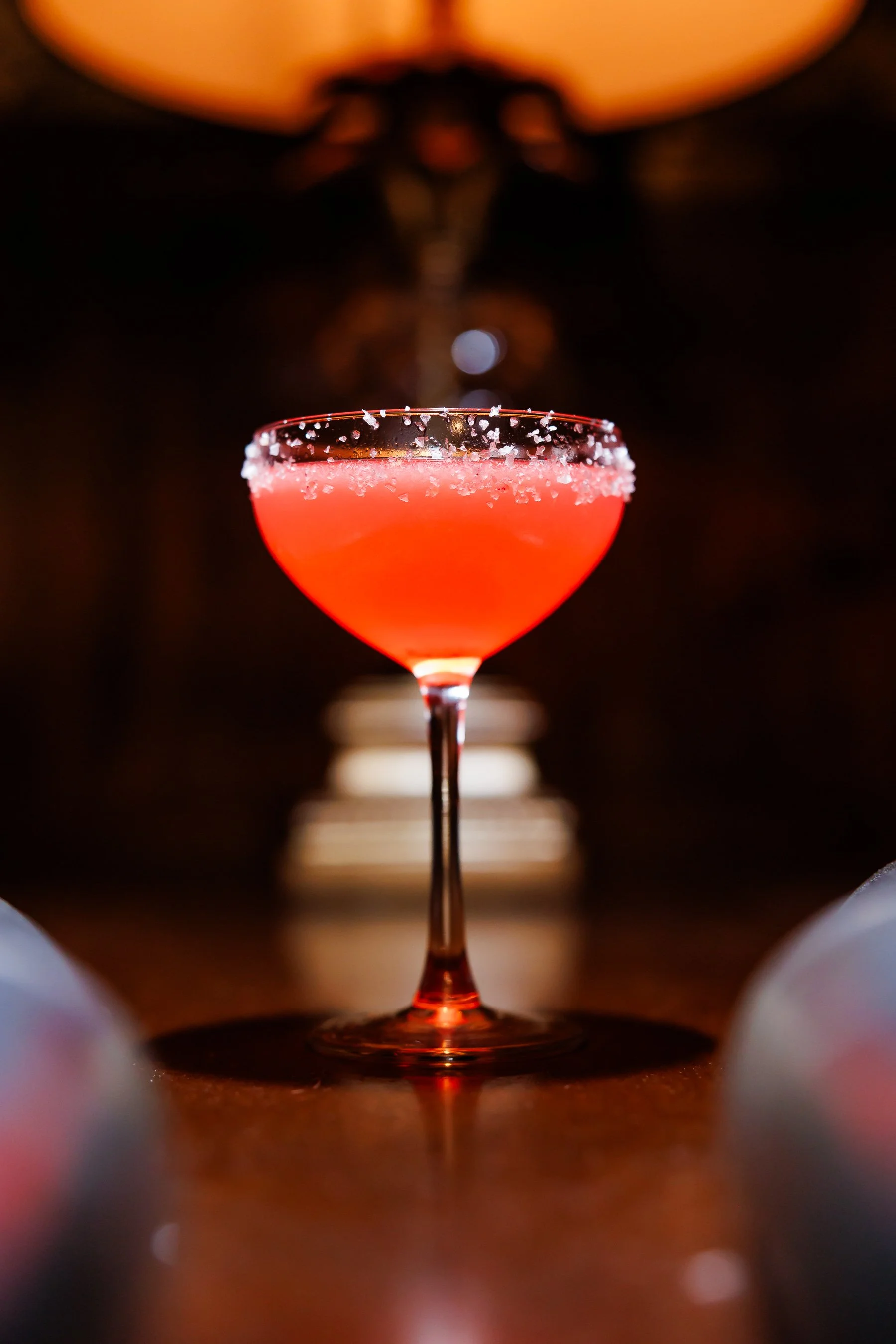 Close-up of a pink cocktail in a coupe glass with salted rim, placed on a wooden surface in dim lighting.