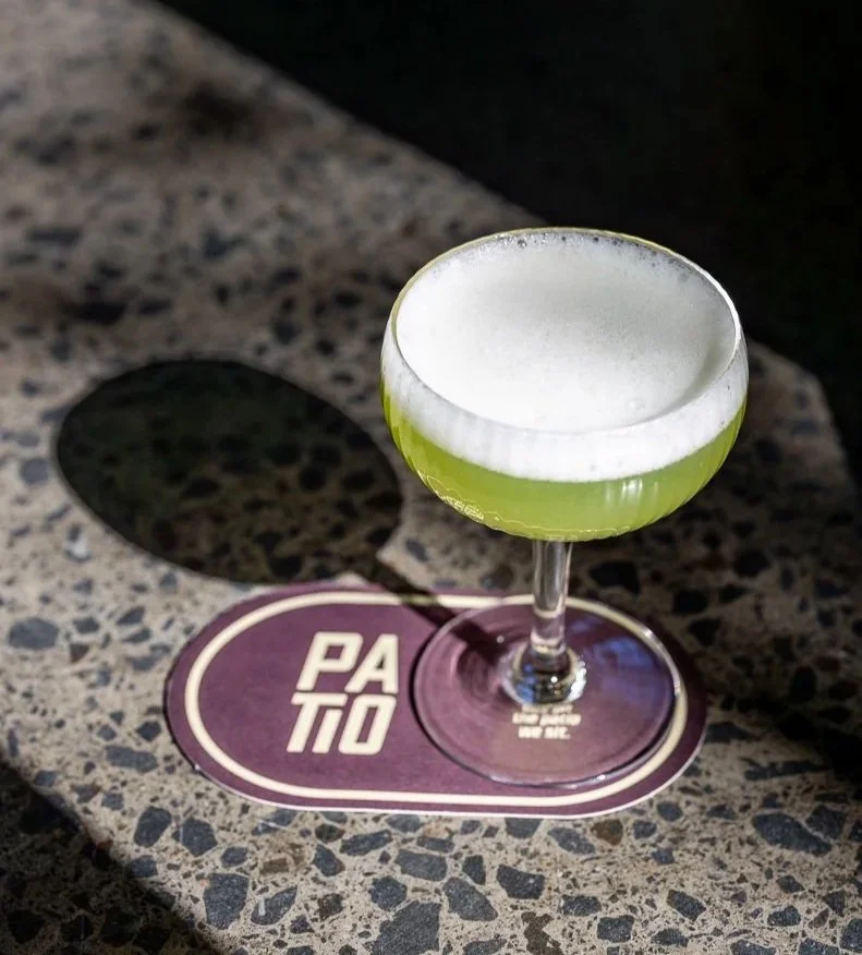 A cocktail glass with a green drink and a frothy white top, placed on a purple and white PASITO coaster, on a speckled gray granite surface.