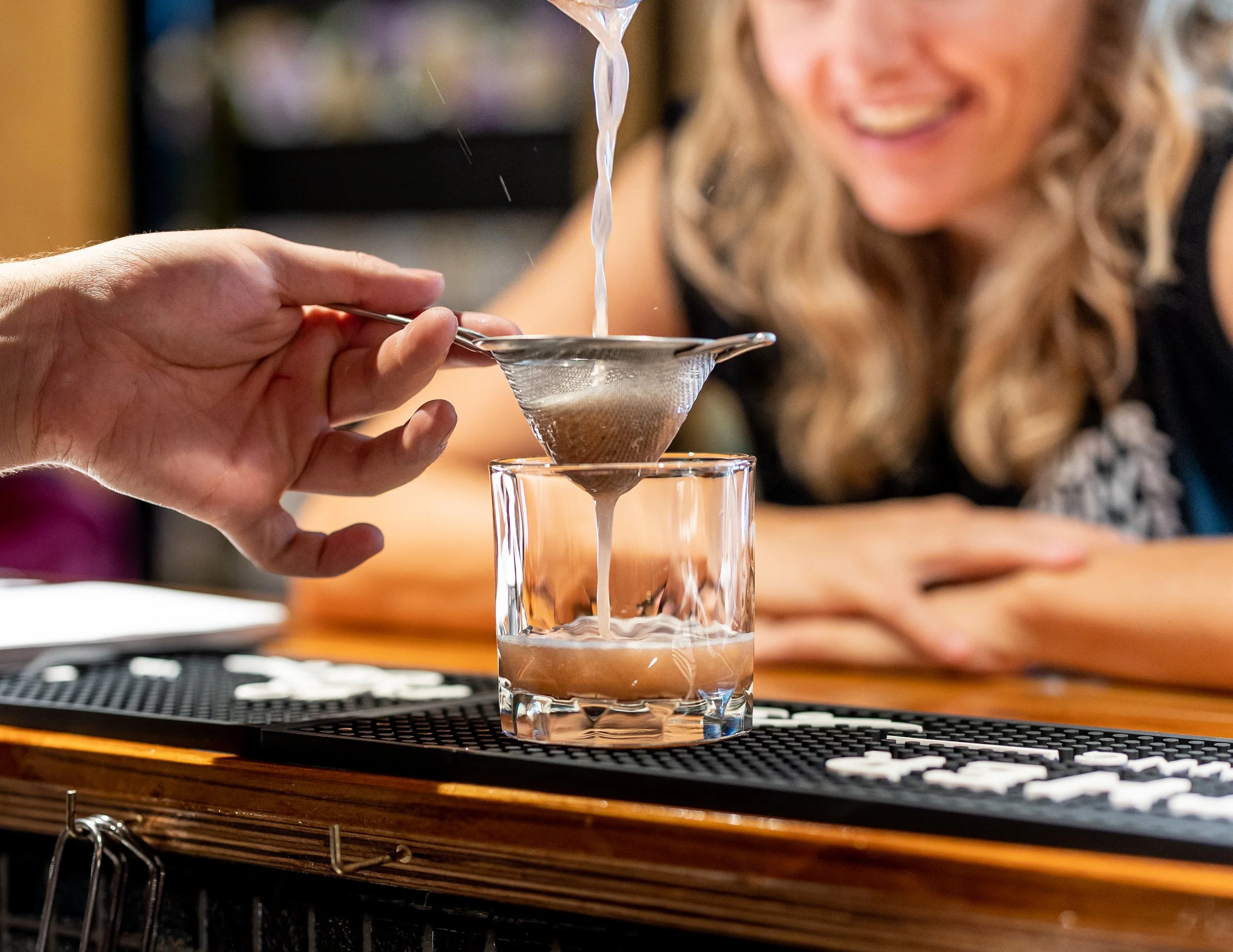 A woman smiling while a person pours a light-colored liquid through a mesh strainer into a glass on a bar counter.