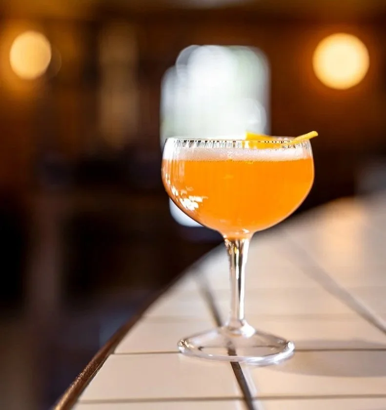 A cocktail glass with a light orange drink, garnished with a lemon peel, on a white table in a dimly lit indoor setting.