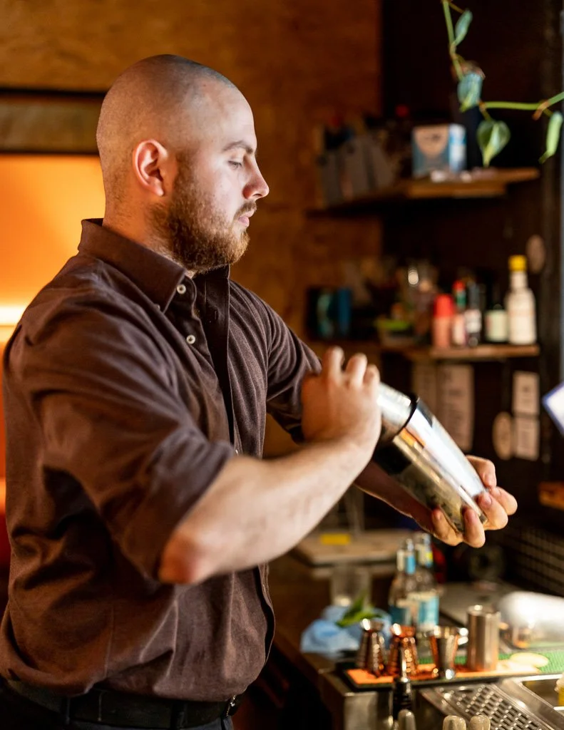 A man with a beard and shaved head preparing a cocktail behind a bar, with various bottles and bar tools in the background.