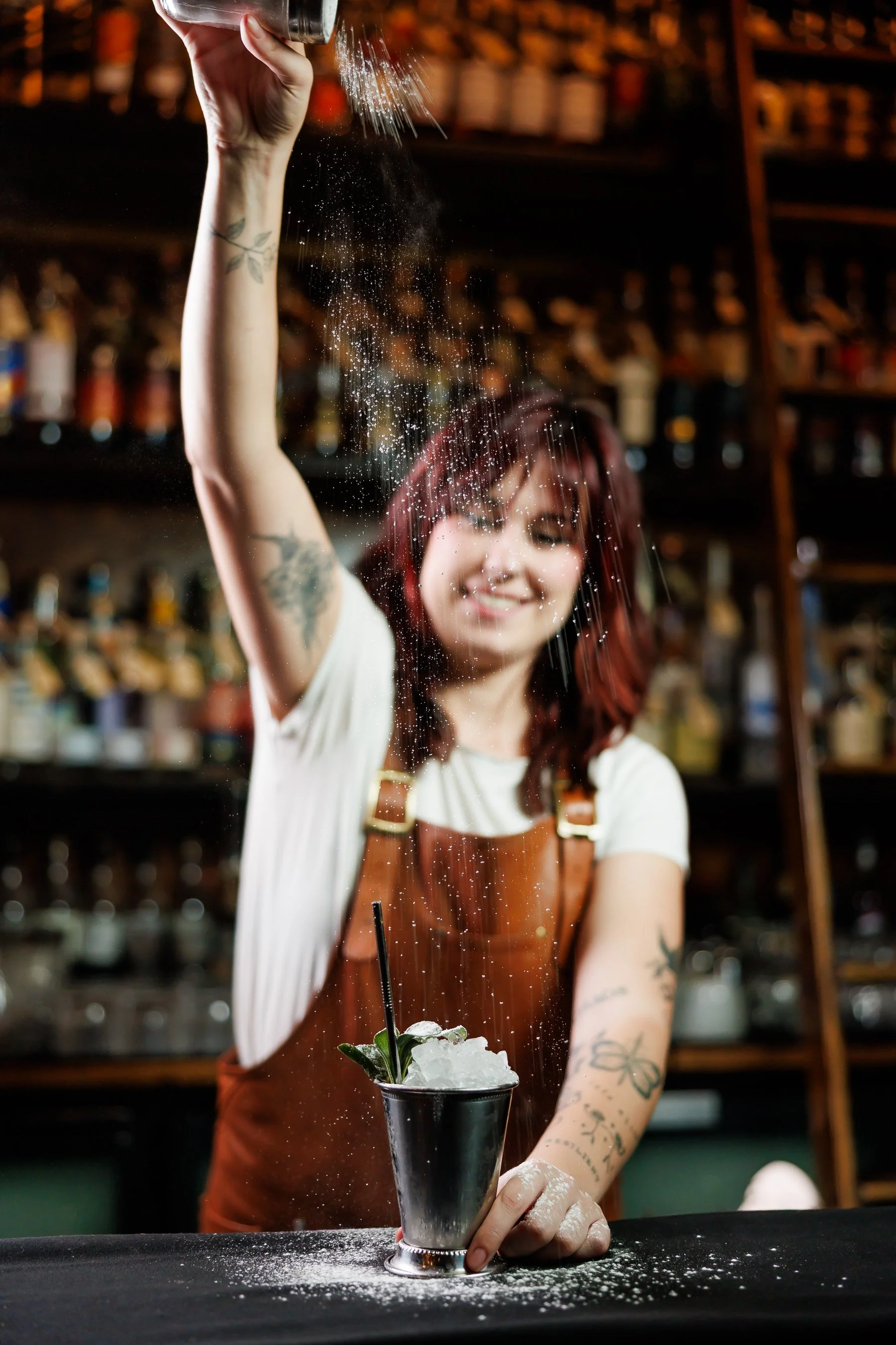 Bartender adding a powdered ingredient to a cocktail with ice, garnished with mint leaves, in a bar setting.
