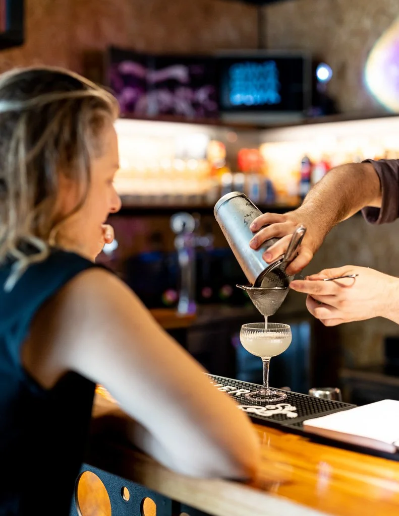 Bartender pouring a cocktail through a strainer into a coupe glass at a bar.