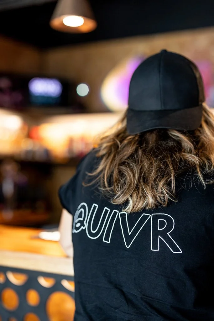 Person with long, wavy hair wearing a black cap and a black T-shirt with the word UIVR on the back, sitting at a bar or counter in a dimly lit setting.