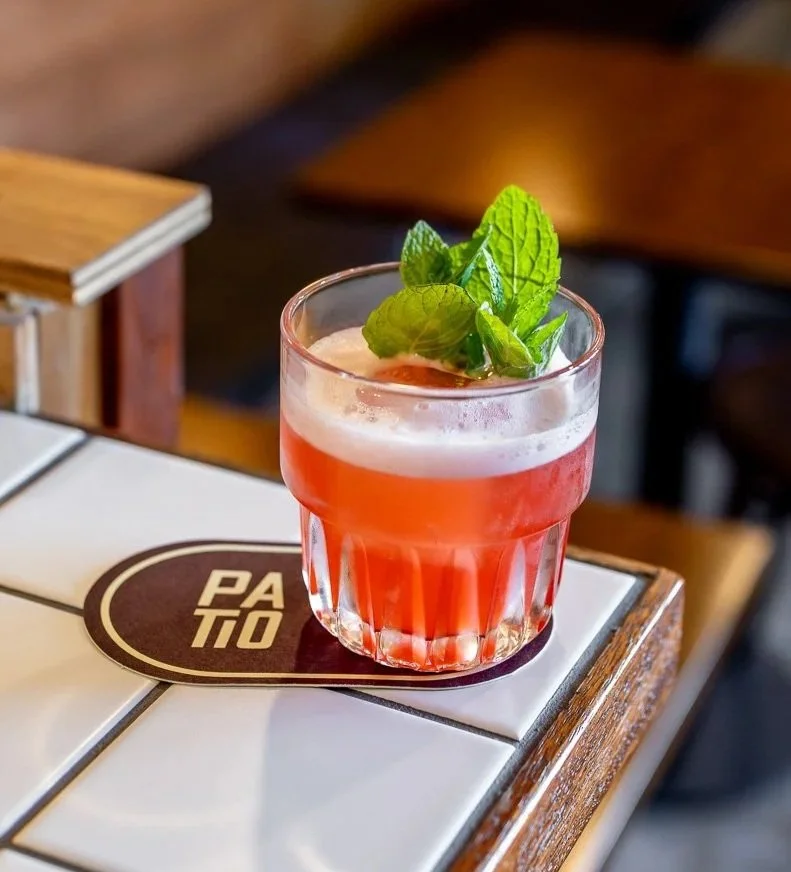 A pink cocktail garnished with fresh mint leaves on a white tiled bar or table with a wooden edge, in a clear glass