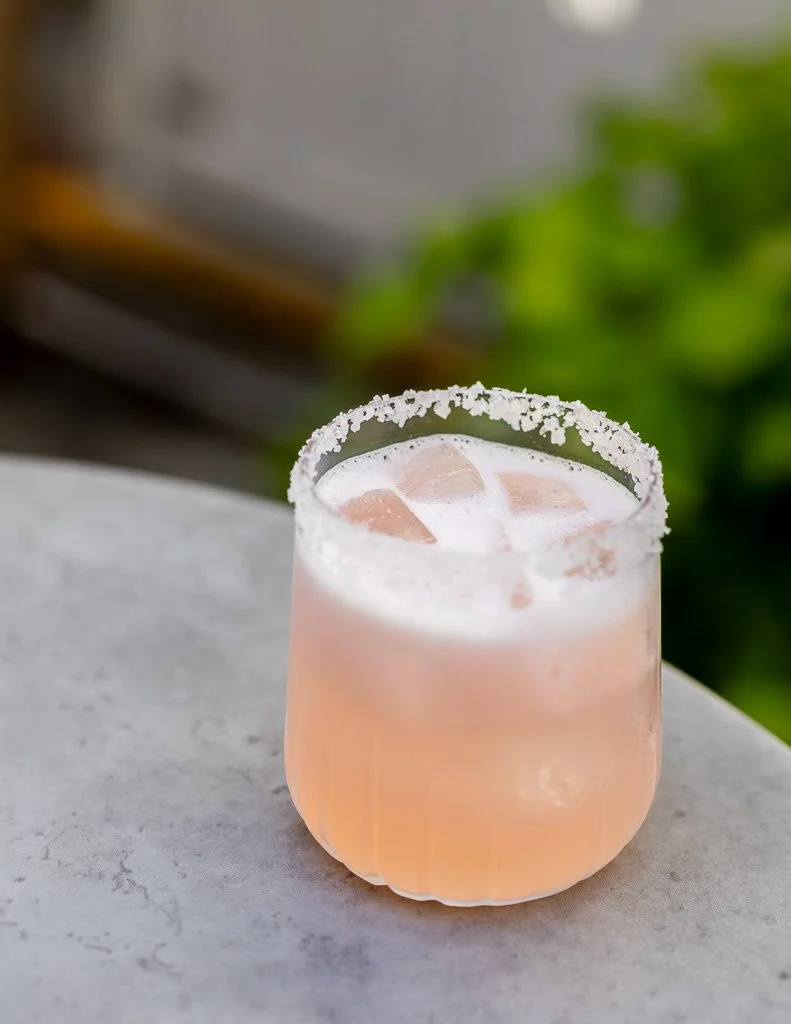 A pink cocktail with ice cubes in a short glass with a salted rim, placed on a concrete surface.