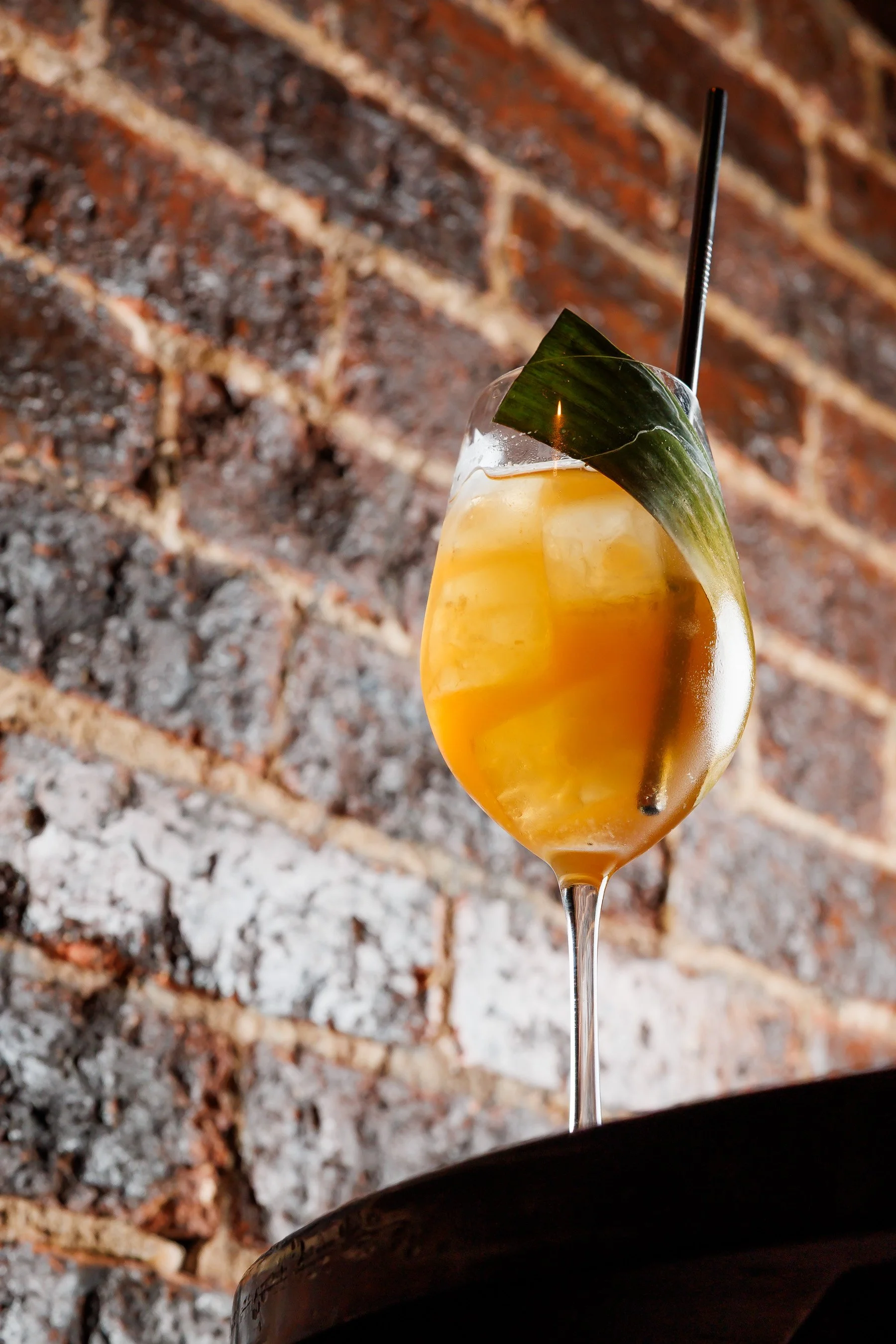 A cocktail glass filled with an orange-colored drink, ice cubes, a black straw, and a green leaf garnish, set against a rustic brick wall background.