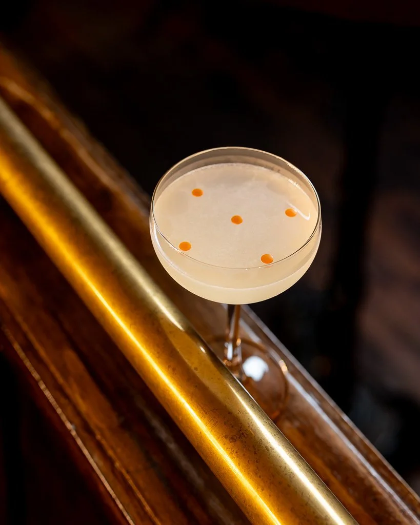 A close-up of a cocktail glass on a wooden bar counter, with a creamy or pale-colored drink garnished with small orange or red dots on top.