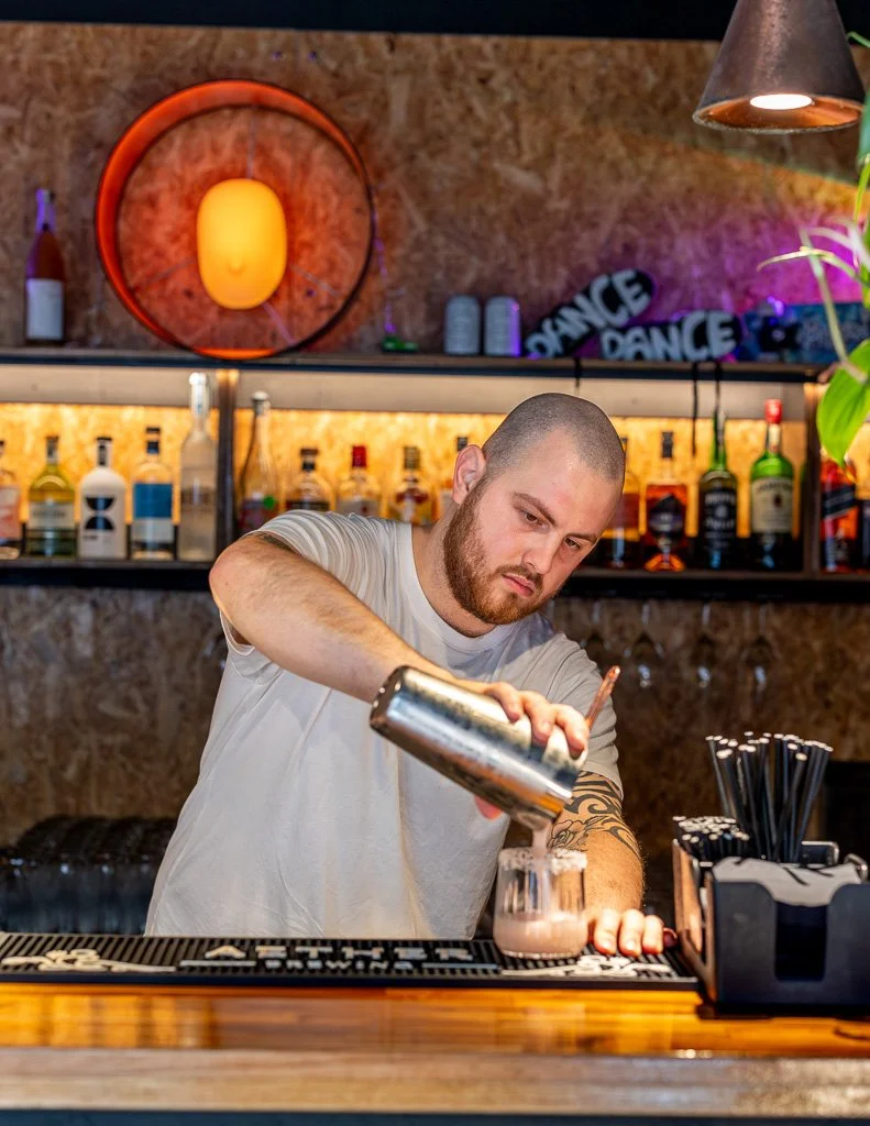 Bartender pouring a drink into a glass at a bar with alcohol bottles in the background.