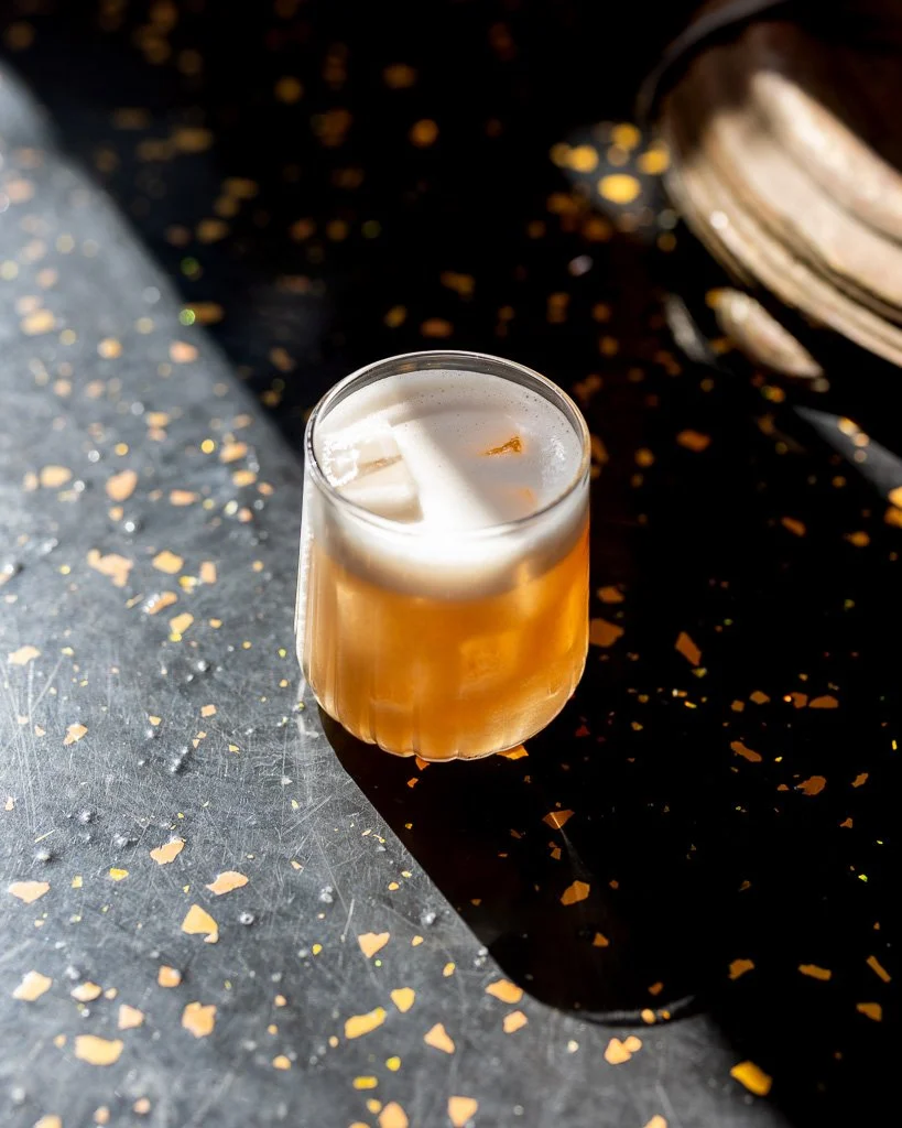 A glass of pale amber beer with a foamy head on a dark surface with scattered gold and orange confetti and a partly visible shoe in the background.