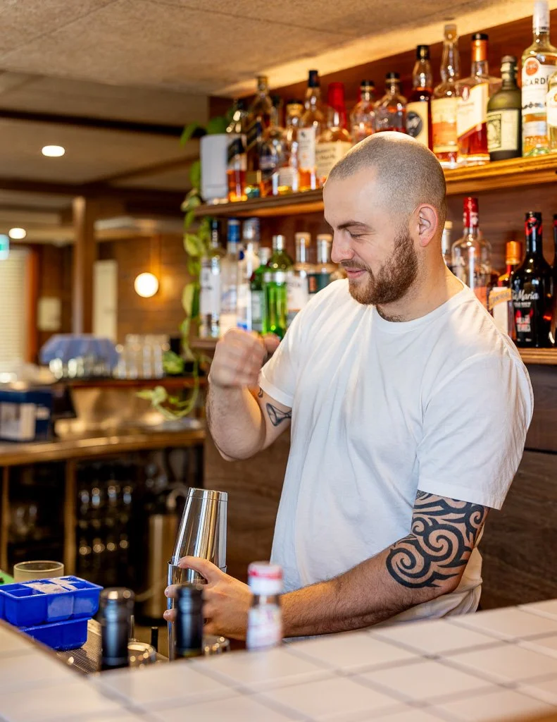 A bartender with a tattooed arm wearing a white t-shirt, smiling and celebrating at the bar, with liquor bottles on shelves in the background.