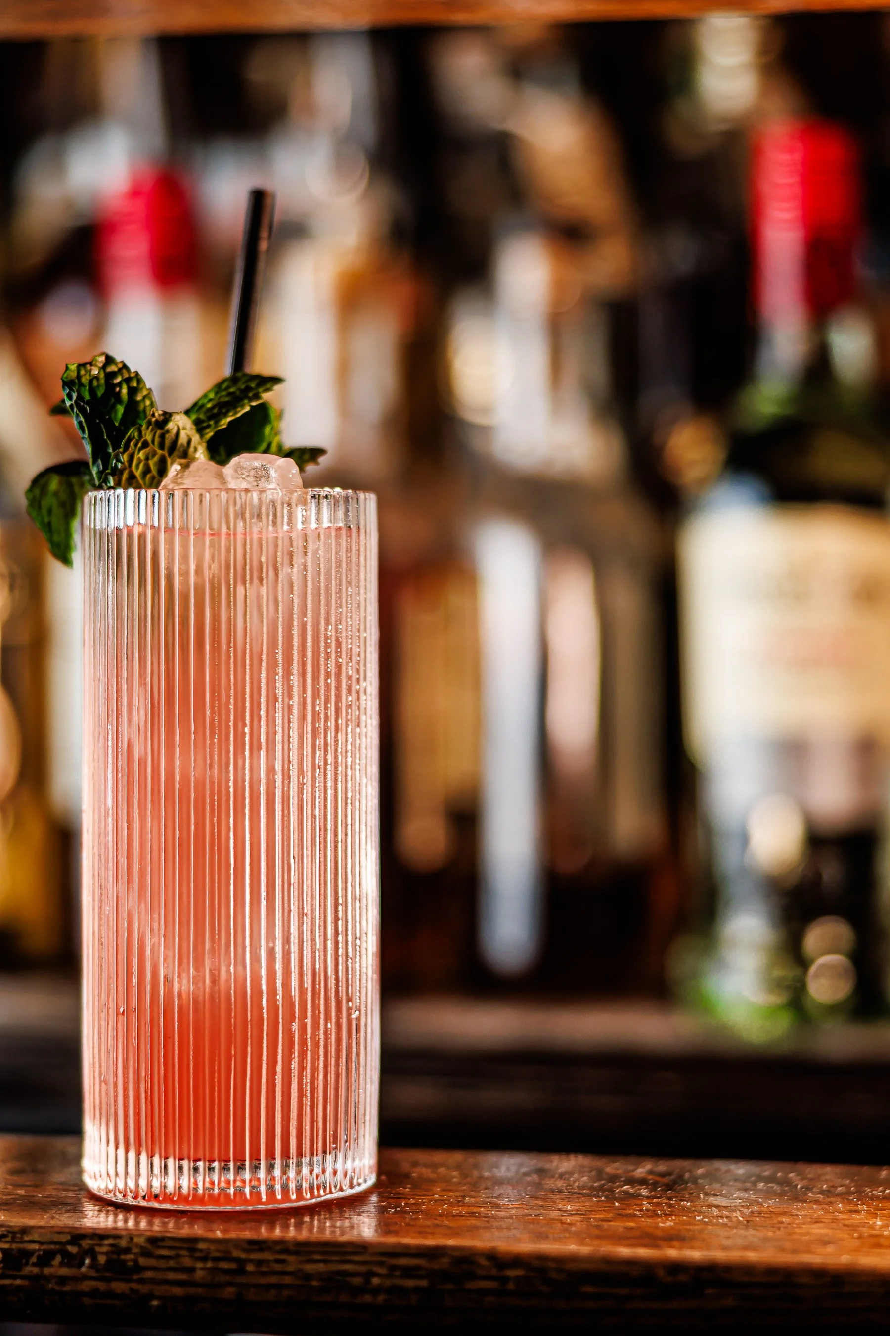A tall glass of pink cocktail with ice, garnished with mint leaves and a black straw, placed on a wooden surface with a blurred bar background.
