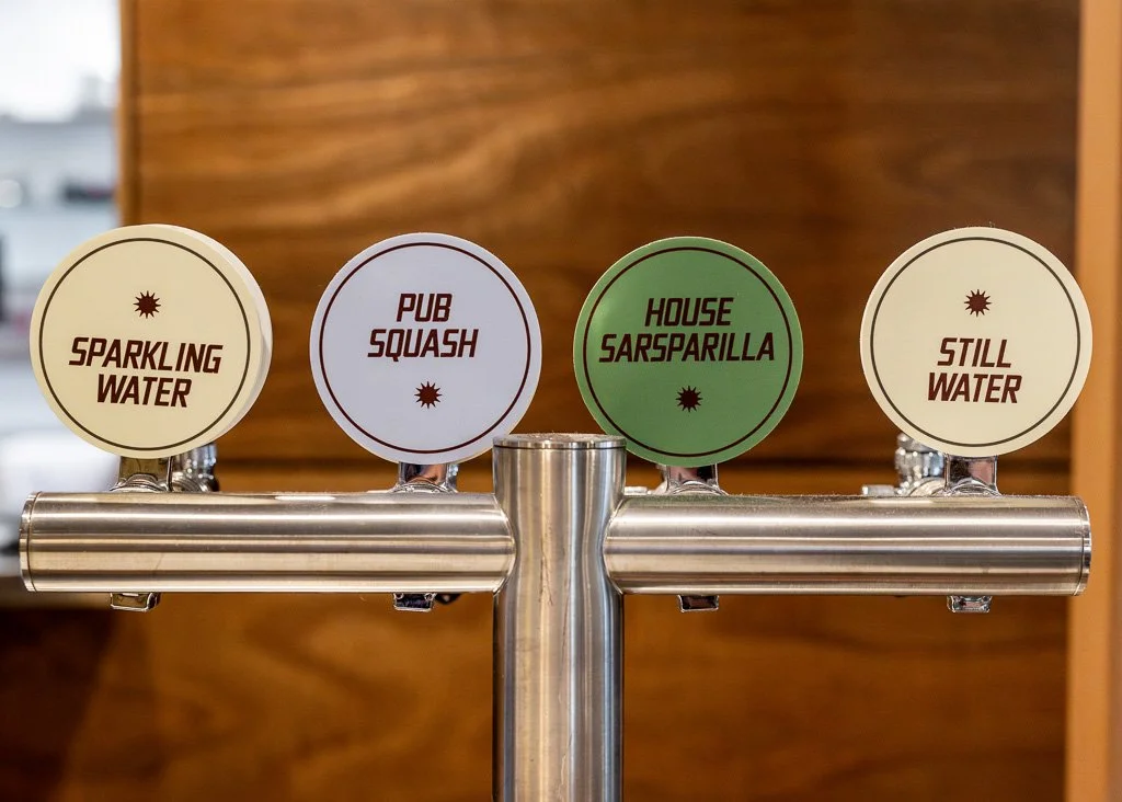 Beer tap handle with four labeled options: Sparkling Water, Pub Squash, House Sarsarilla, Still Water, mounted on a metal pipe against a wooden background.