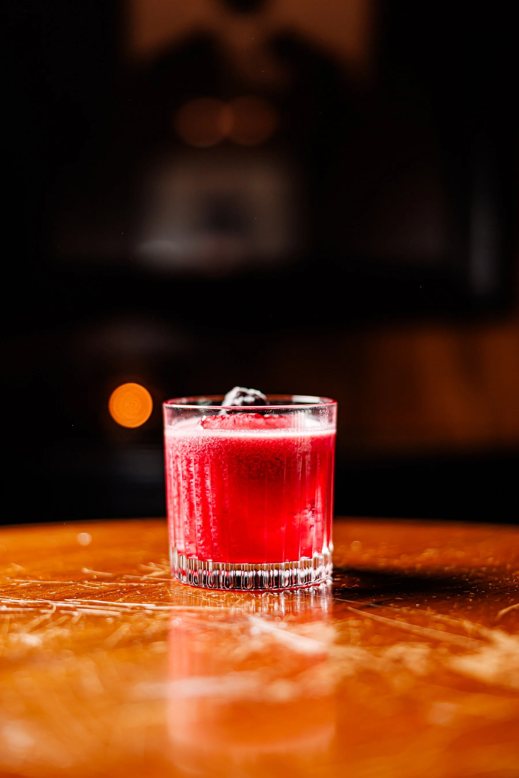 A close-up of a red cocktail with ice in a clear glass on a wooden surface, with a dark blurred background.