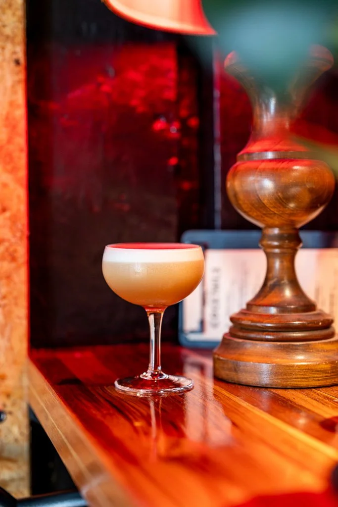 Cream-colored cocktail in a coupe glass on a wooden surface with a tall wooden sculpture in the background.