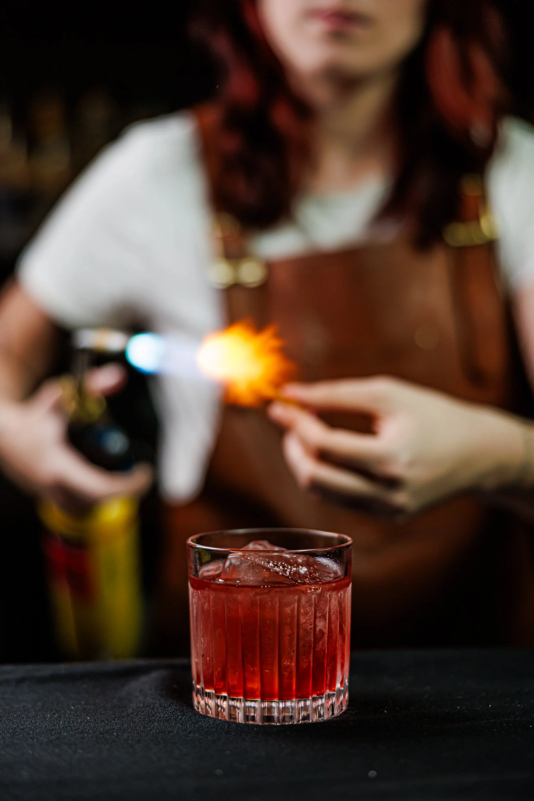 A red cocktail with ice cubes in a glass on a black surface, with a person in the background holding a sparks-generating device near their chest.